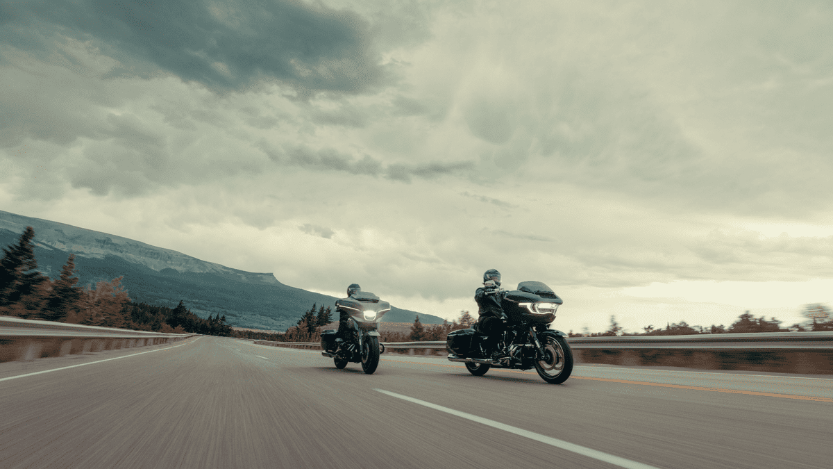 Two 2025 Harley-Davidson® Grand American Touring bikes riding side-by-side on a highway with a mountain view, designed for long-distance comfort and power.