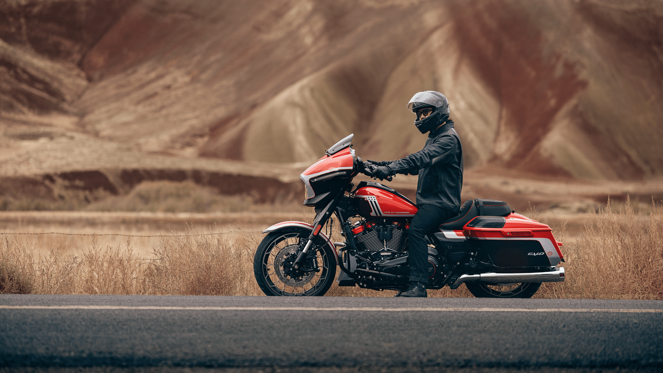 A person wearing a helmet and riding gear sits on a red Harley-Davidson CVO™ Street Glide® motorcycle on a road with a mountainous background.