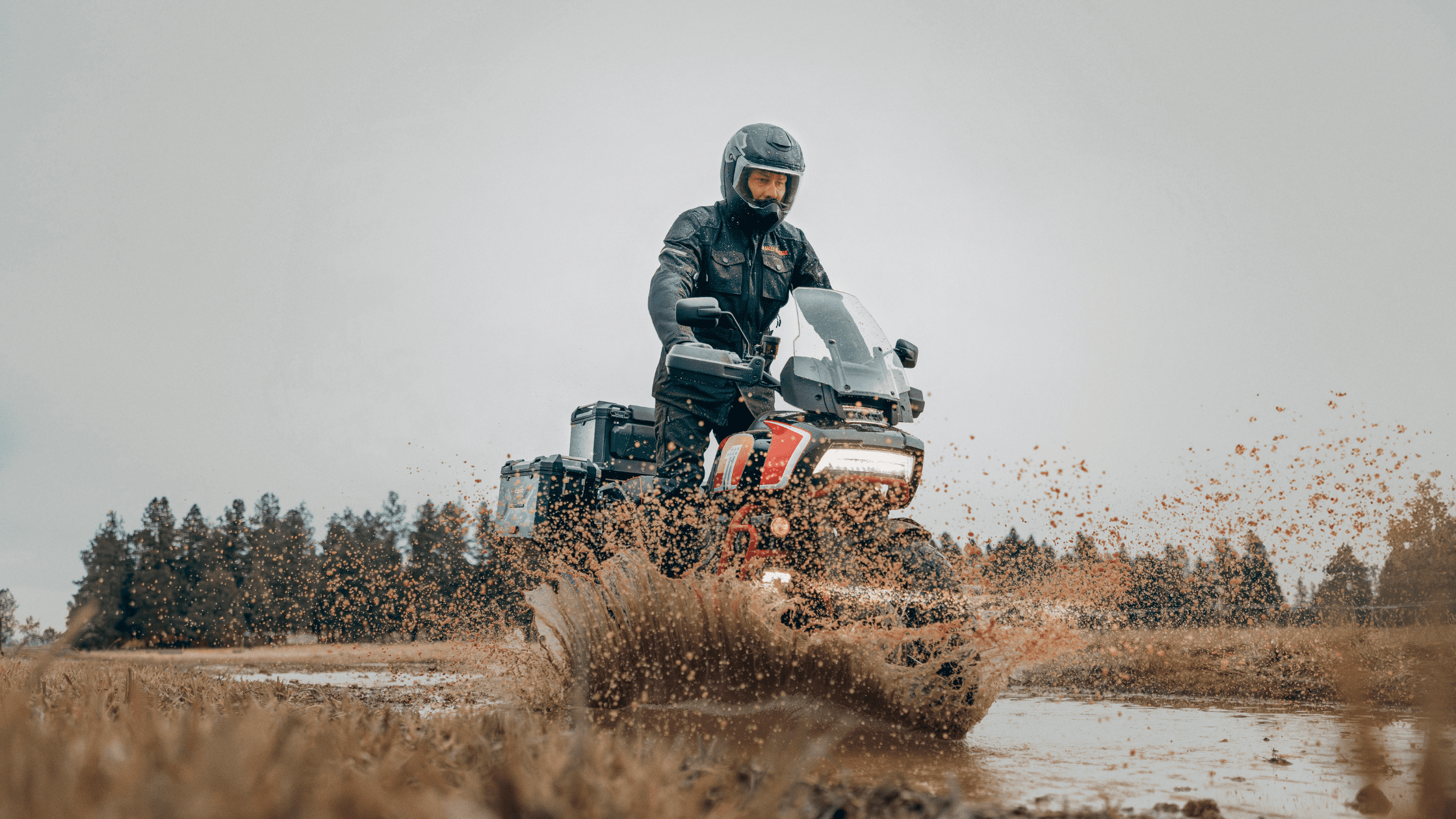 A rider on a Harley-Davidson CVO™ Pan America® motorcycle splashing through a muddy field.