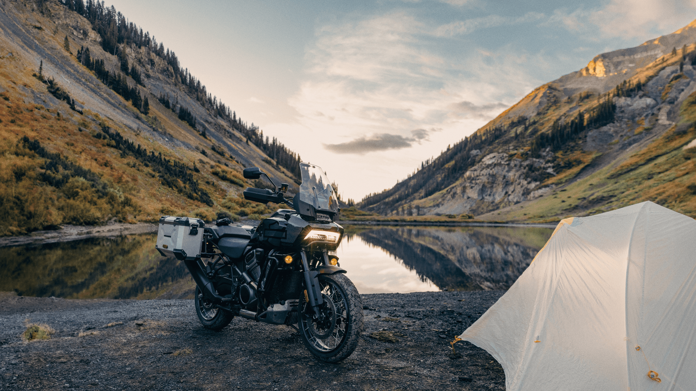 A black motorcycle is parked beside a tent near a serene lake, surrounded by mountains with lush greenery. The sun is setting, casting a warm glow on the landscape.