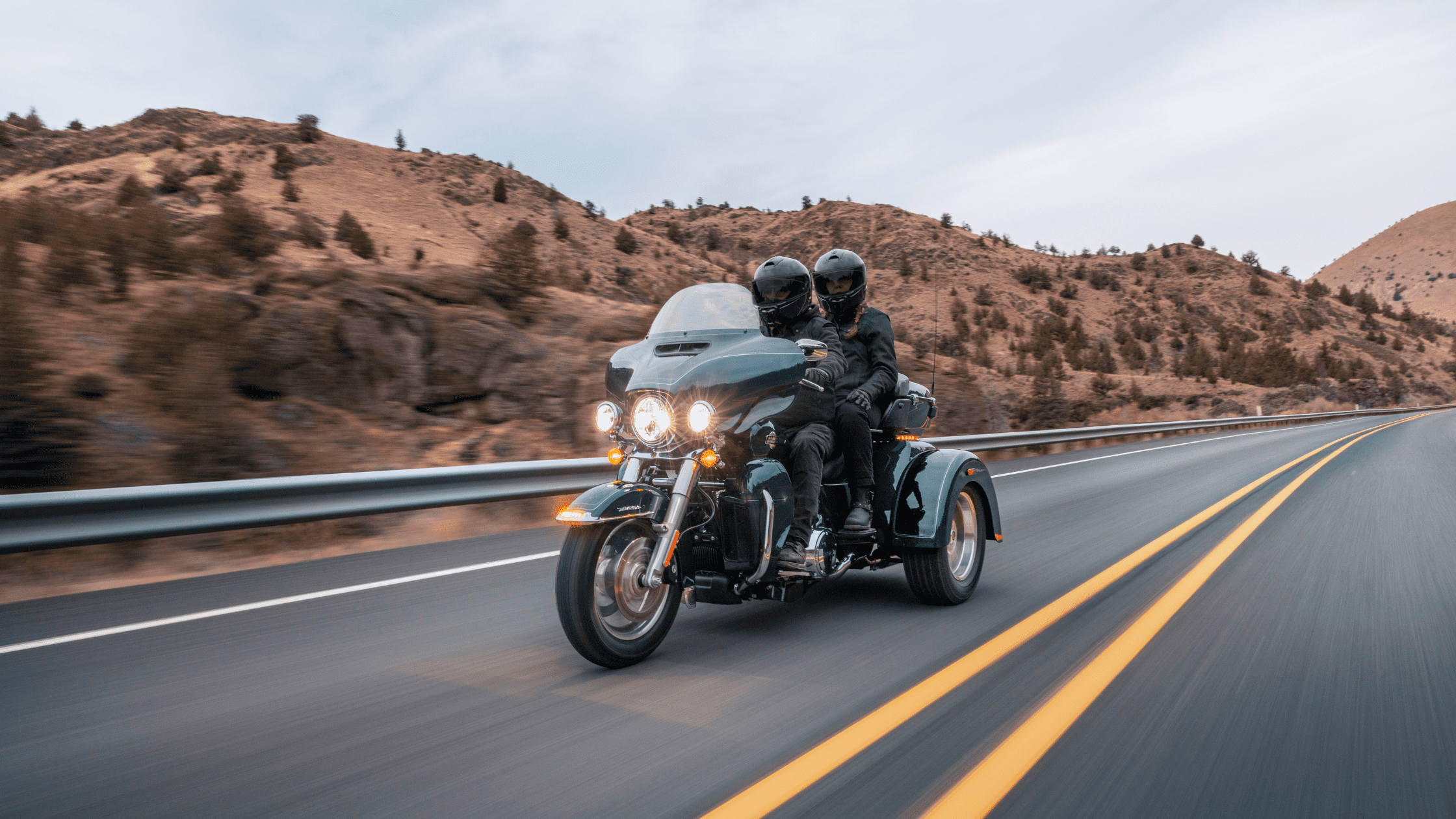 A couple riding a Harley-Davidson Tri Glide Ultra motorcycle on a scenic highway through a mountainous landscape.