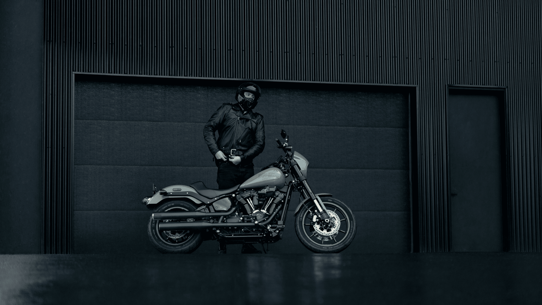 Person in a leather jacket and helmet standing next to a Harley-Davidson motorcycle in front of a modern, dark-colored building.