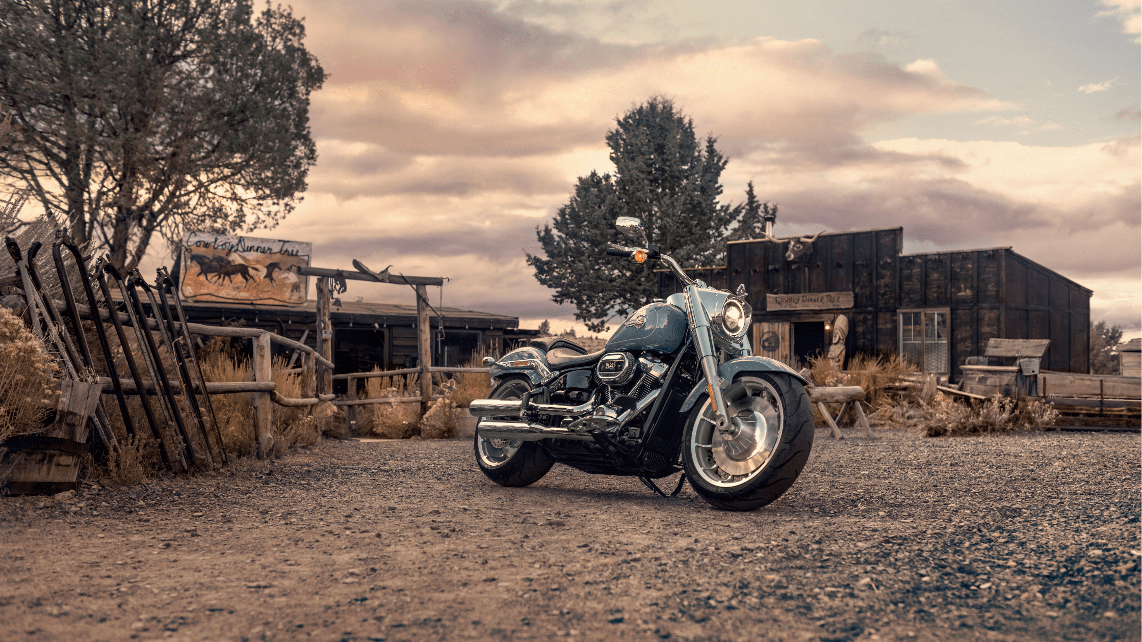 Harley-Davidson Fat Boy® 114 motorcycle parked in front of a rustic cowboy-themed setting at sunset.