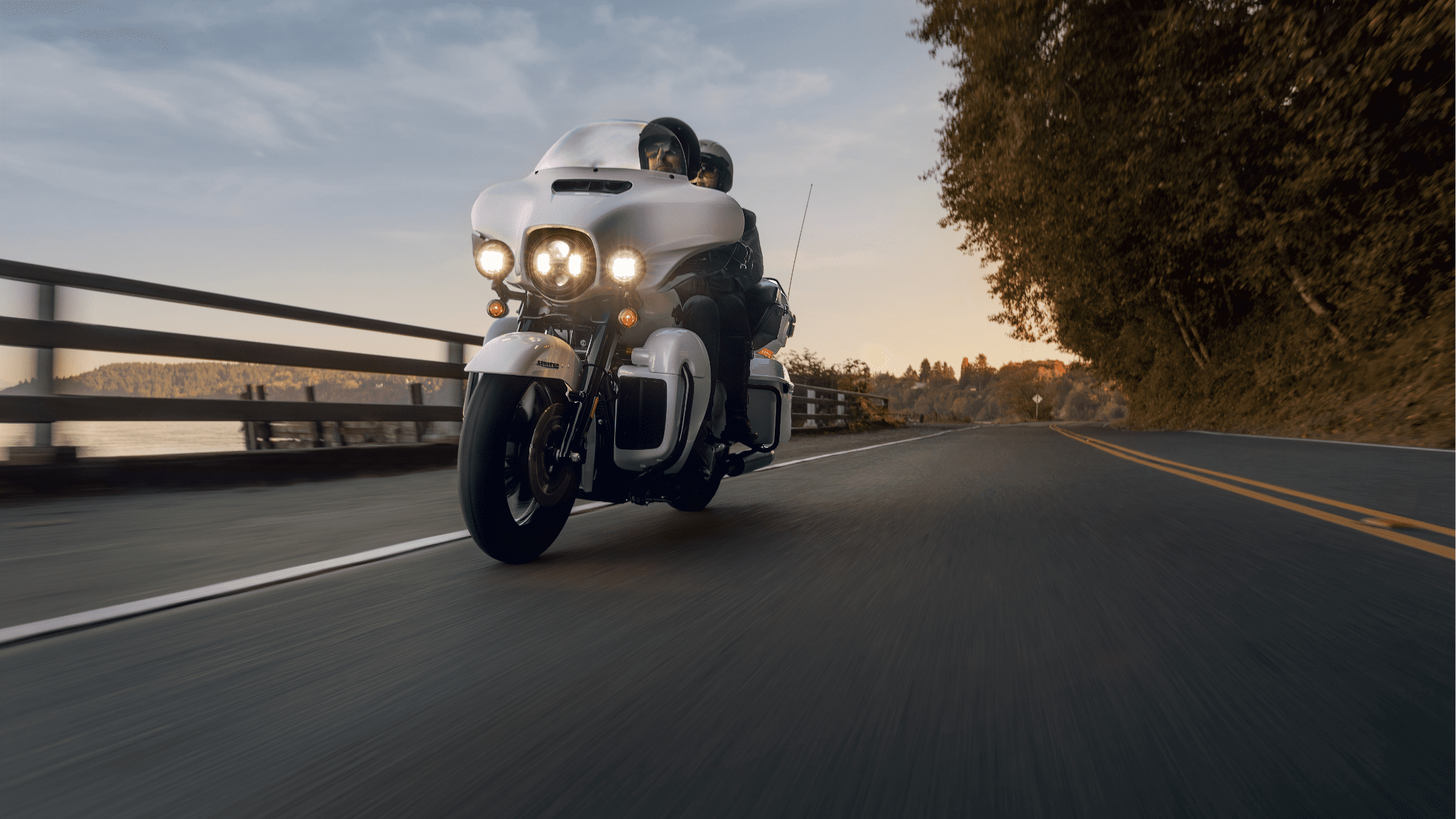 Harley-Davidson Ultra Limited motorcycle riding on a road beside a body of water at sunset.