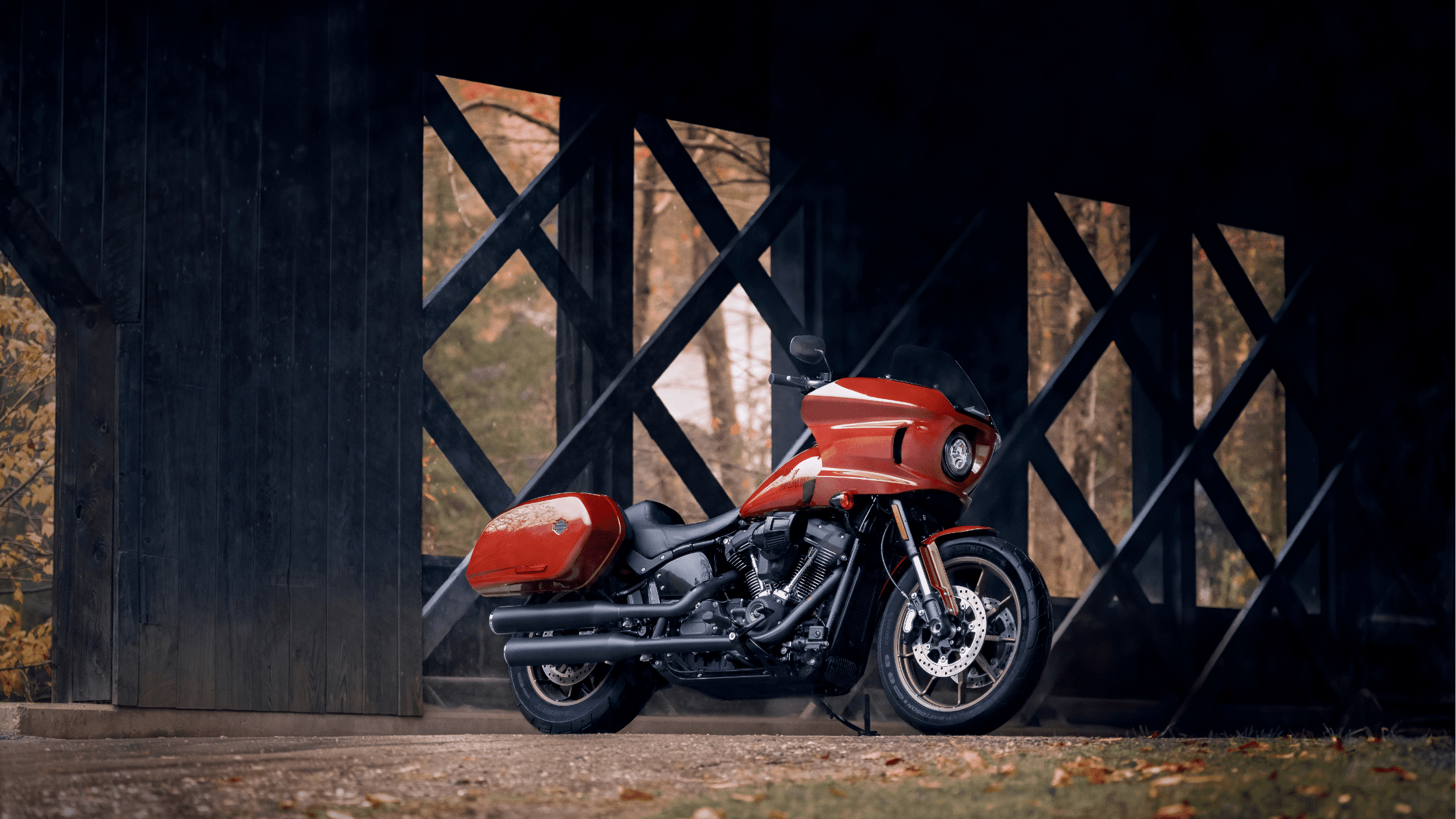 Red 2024 Harley-Davidson® Low Rider® ST parked in front of a rustic wooden bridge with an autumnal backdrop.