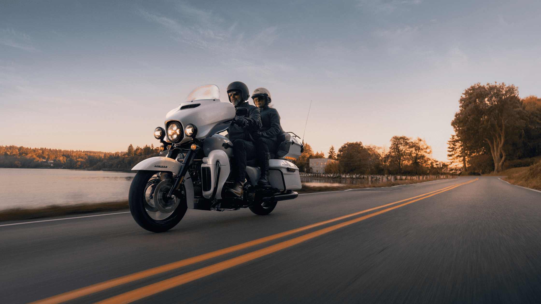 Two riders on a Harley-Davidson Ultra Limited motorcycle cruising down a scenic road at sunset
