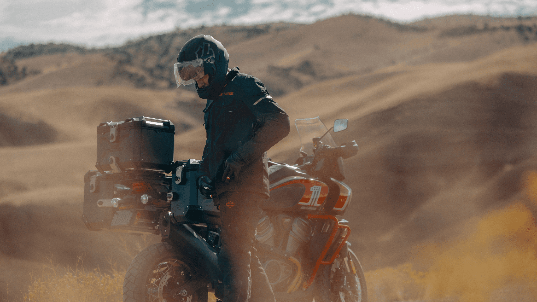 A motorcyclist wearing protective gear stands next to a fully equipped adventure motorcycle in a rugged, mountainous landscape, with dusty trails and rolling hills in the background.