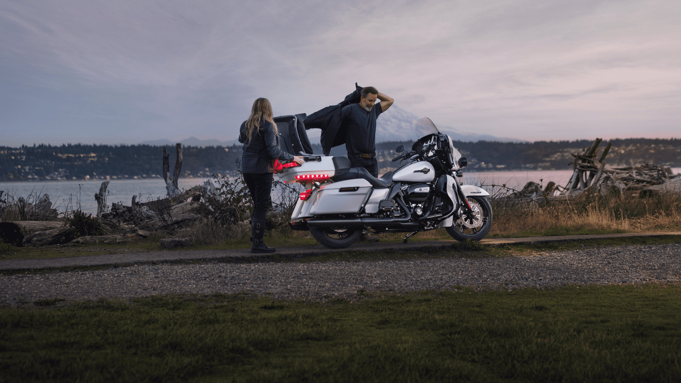 A 2024 Harley-Davidson® Ultra Limited® parked by a lakeside, with a couple standing nearby, one person adjusting their jacket while the other rests a hand on the bike's rear.