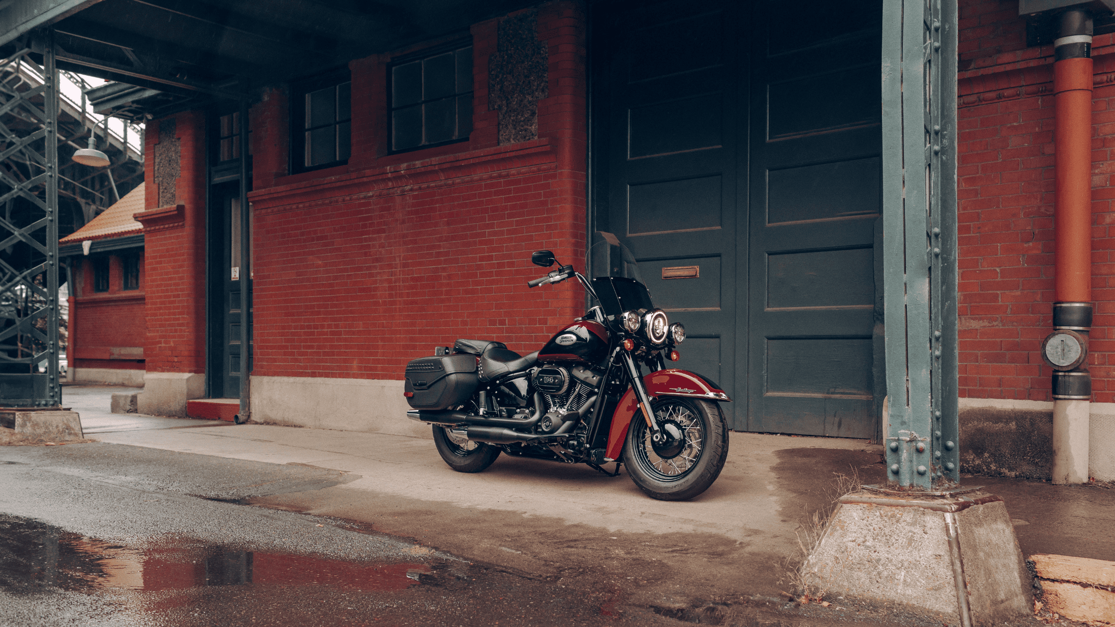 A 2024 Harley-Davidson® Heritage Classic motorcycle parked in front of a red brick building under an overpass.
