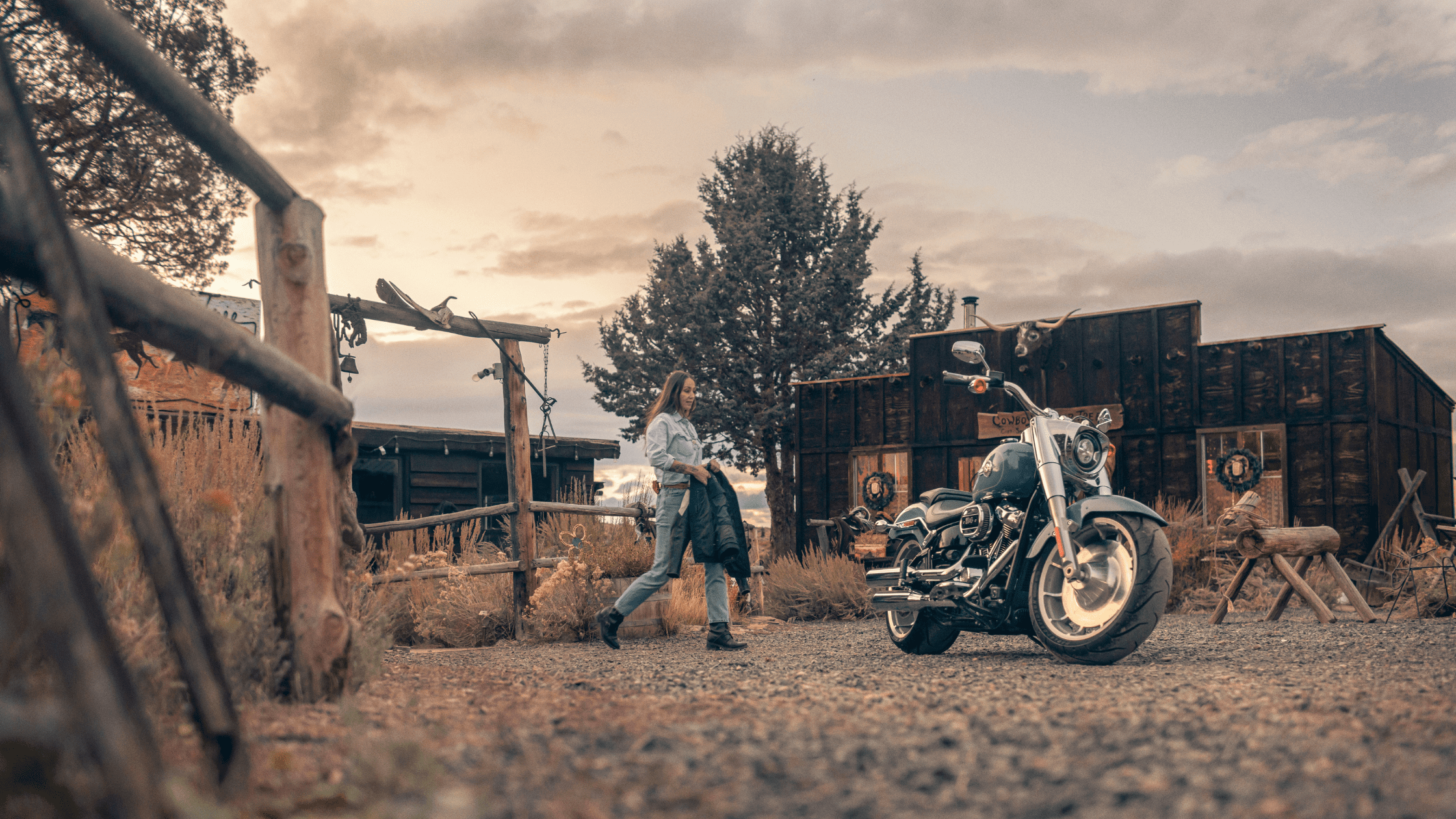 A 2024 Harley-Davidson® Fat Boy® 114 parked outside a rustic wooden building, with a woman in the background walking towards it, holding a jacket.