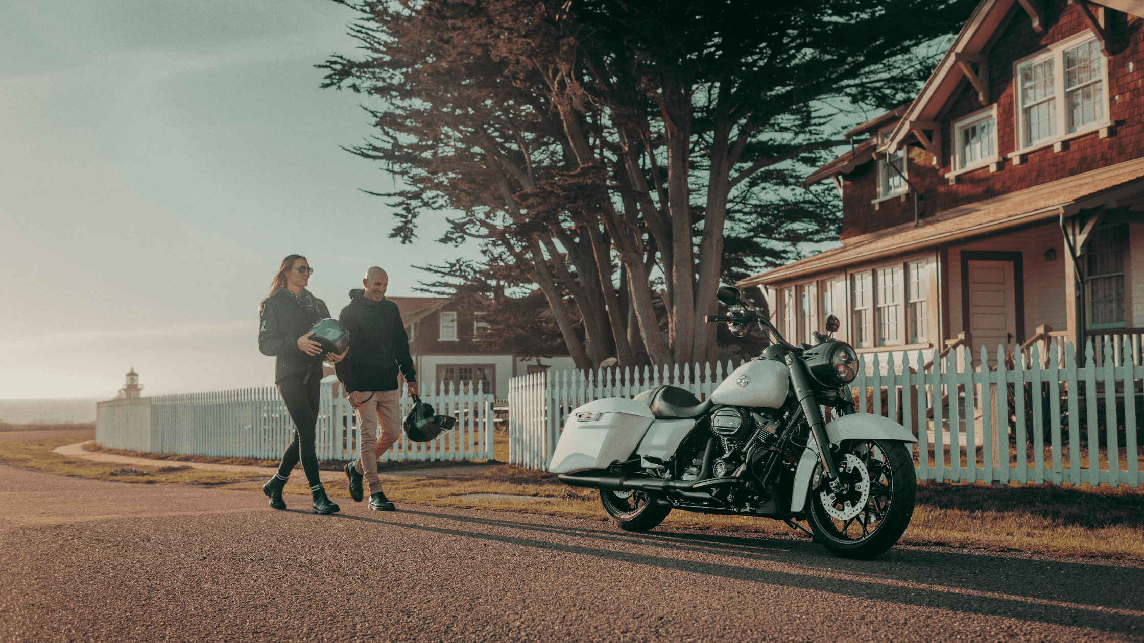 A couple walks past a parked 2024 Harley-Davidson® Road King® Special motorcycle near a picket fence in a serene neighborhood.