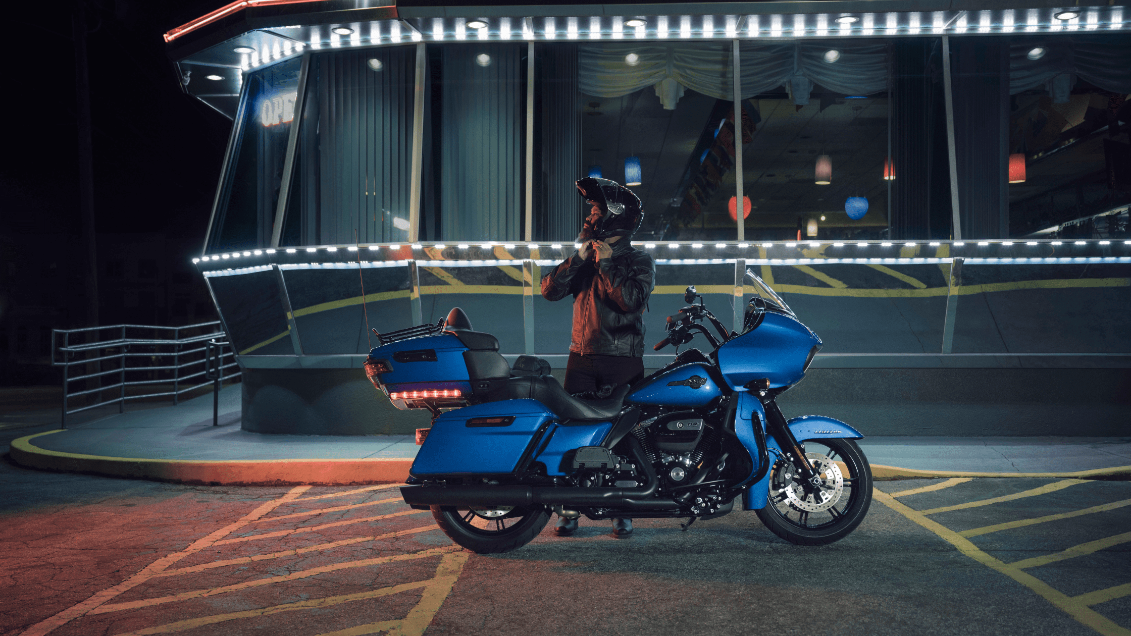 A rider stands next to a blue Harley-Davidson® Road Glide® Limited motorcycle at night, in front of a brightly lit diner.