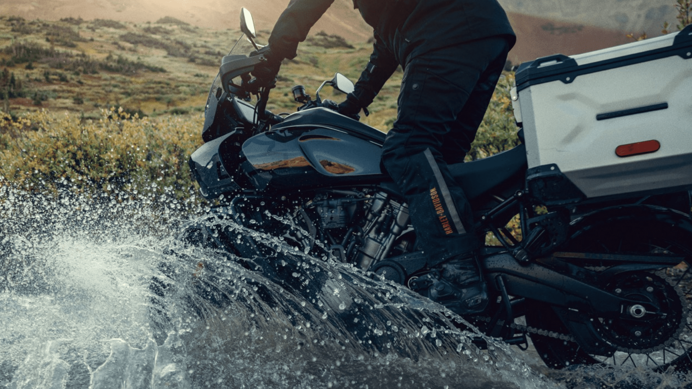 A rider on a 2024 Harley-Davidson® Pan America® 1250 Special crossing a shallow stream, splashing water while navigating off-road terrain