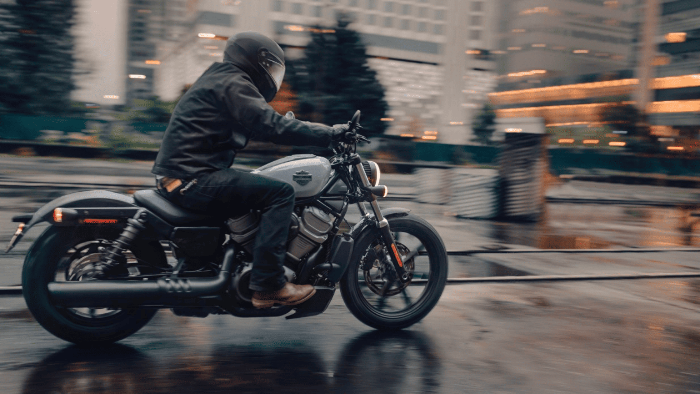 Motorcyclist riding a Harley-Davidson Nightster® through a wet city street, with blurred urban lights and reflections on the road.