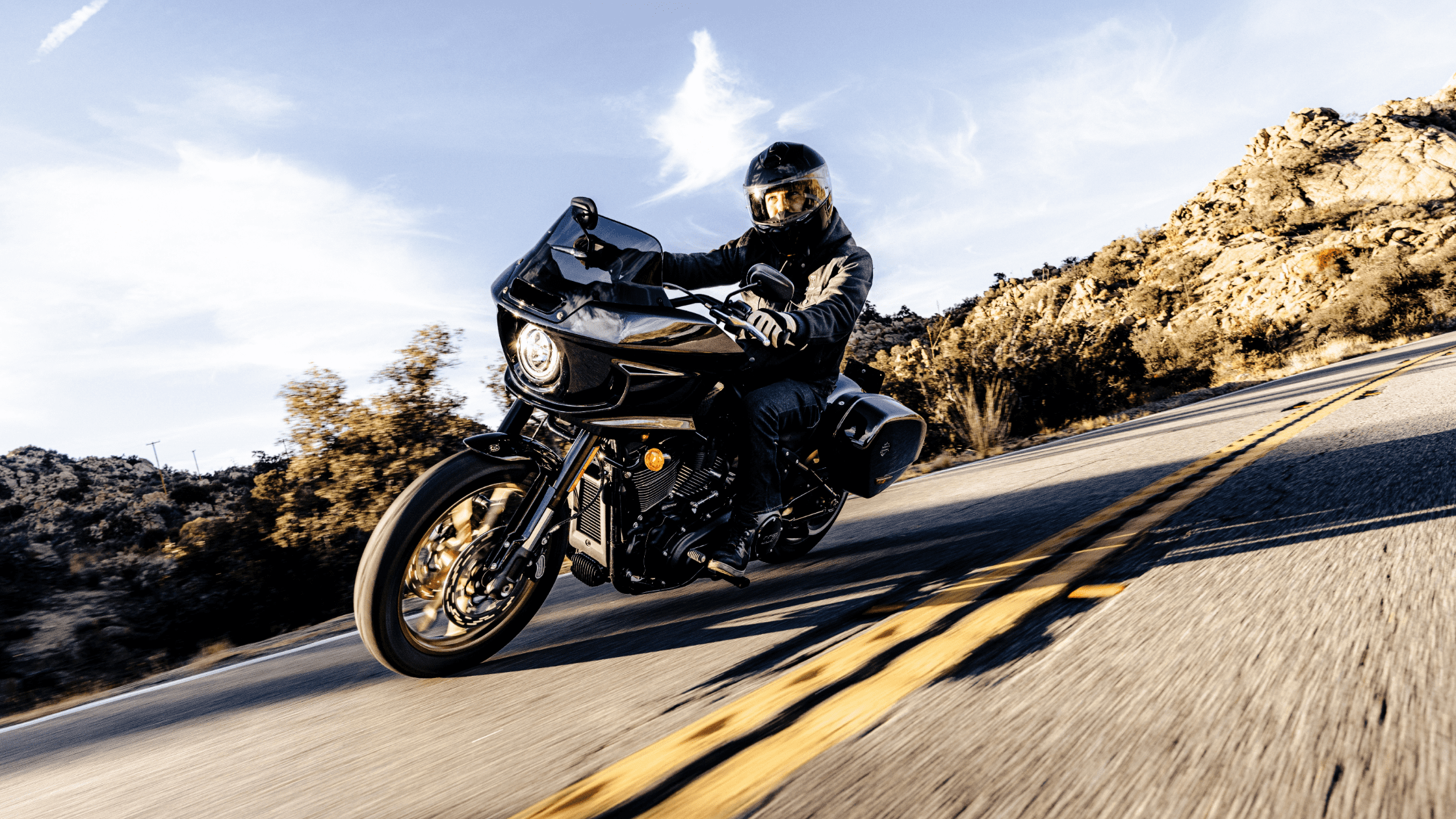 A rider leaning into a turn on a 2024 Harley-Davidson® Low Rider® ST, cruising down a scenic desert road with mountains in the background