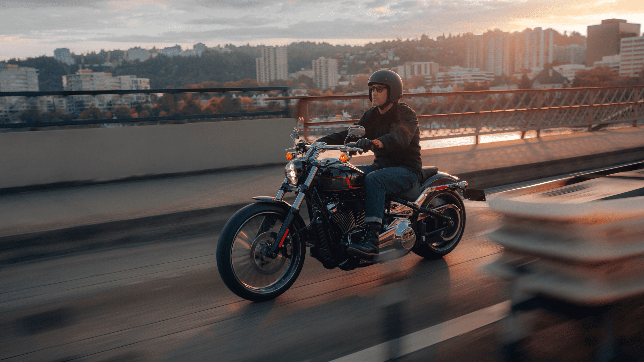 A rider on a 2024 Harley-Davidson® Breakout® 117 cruising across a city bridge at sunset with buildings and hills in the background