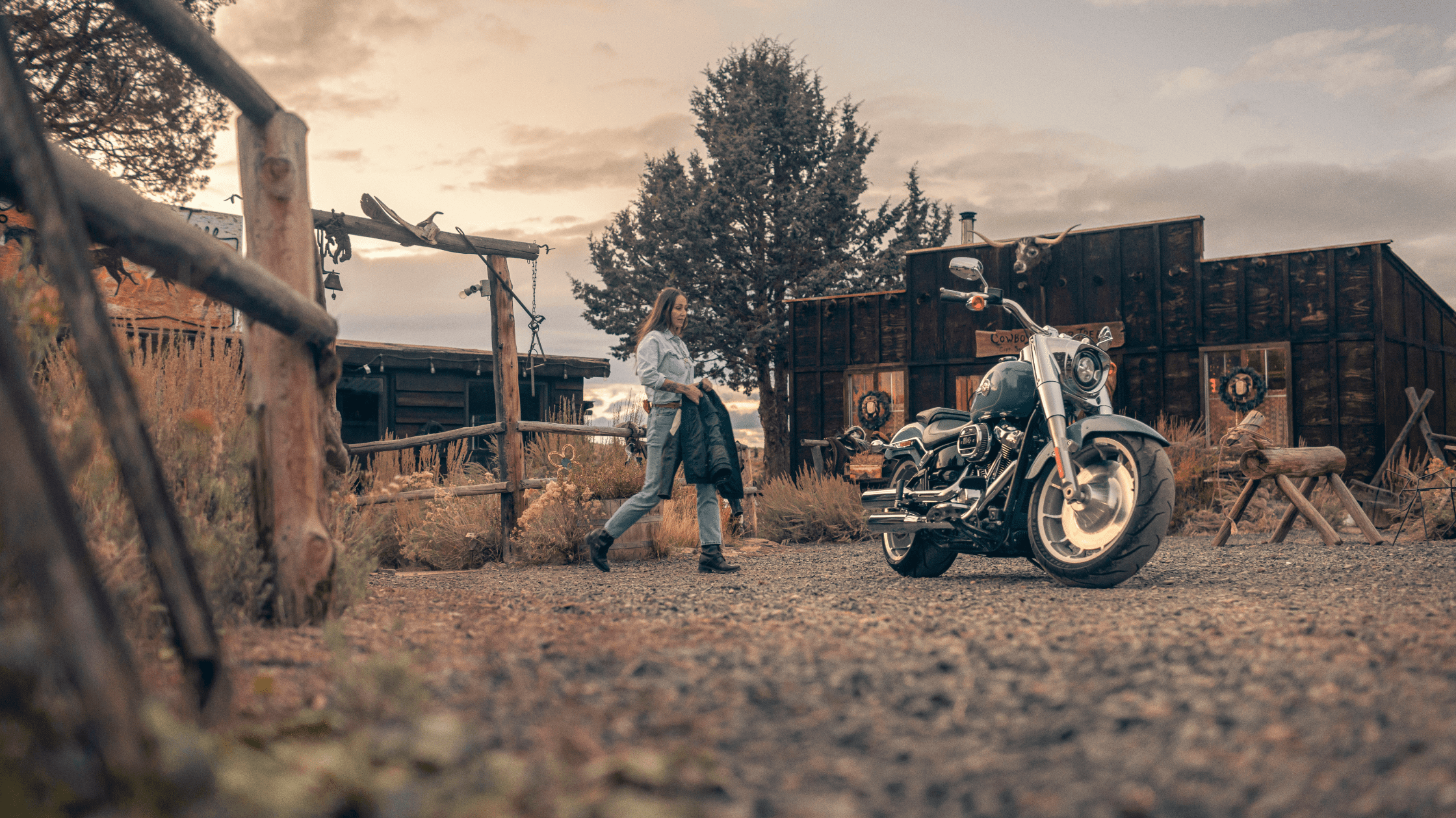 A 2024 Harley-Davidson® Fat Boy® 114 parked in front of a rustic cabin as a woman walks nearby