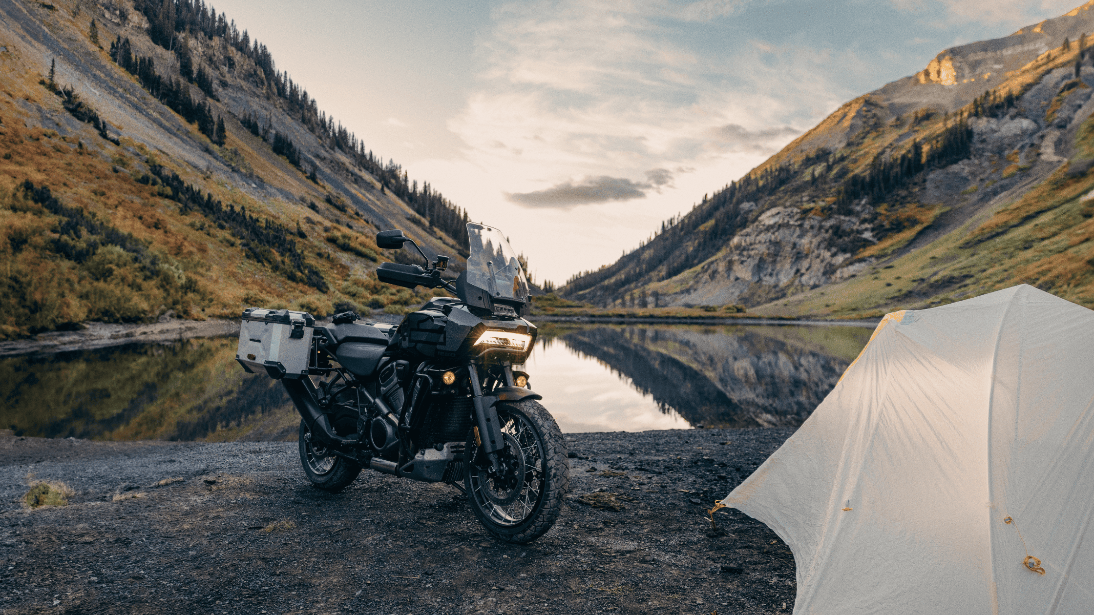 Harley-Davidson Pan America® 1250 Special parked next to a tent by a mountain lake, with stunning valley and mountain scenery at sunset.