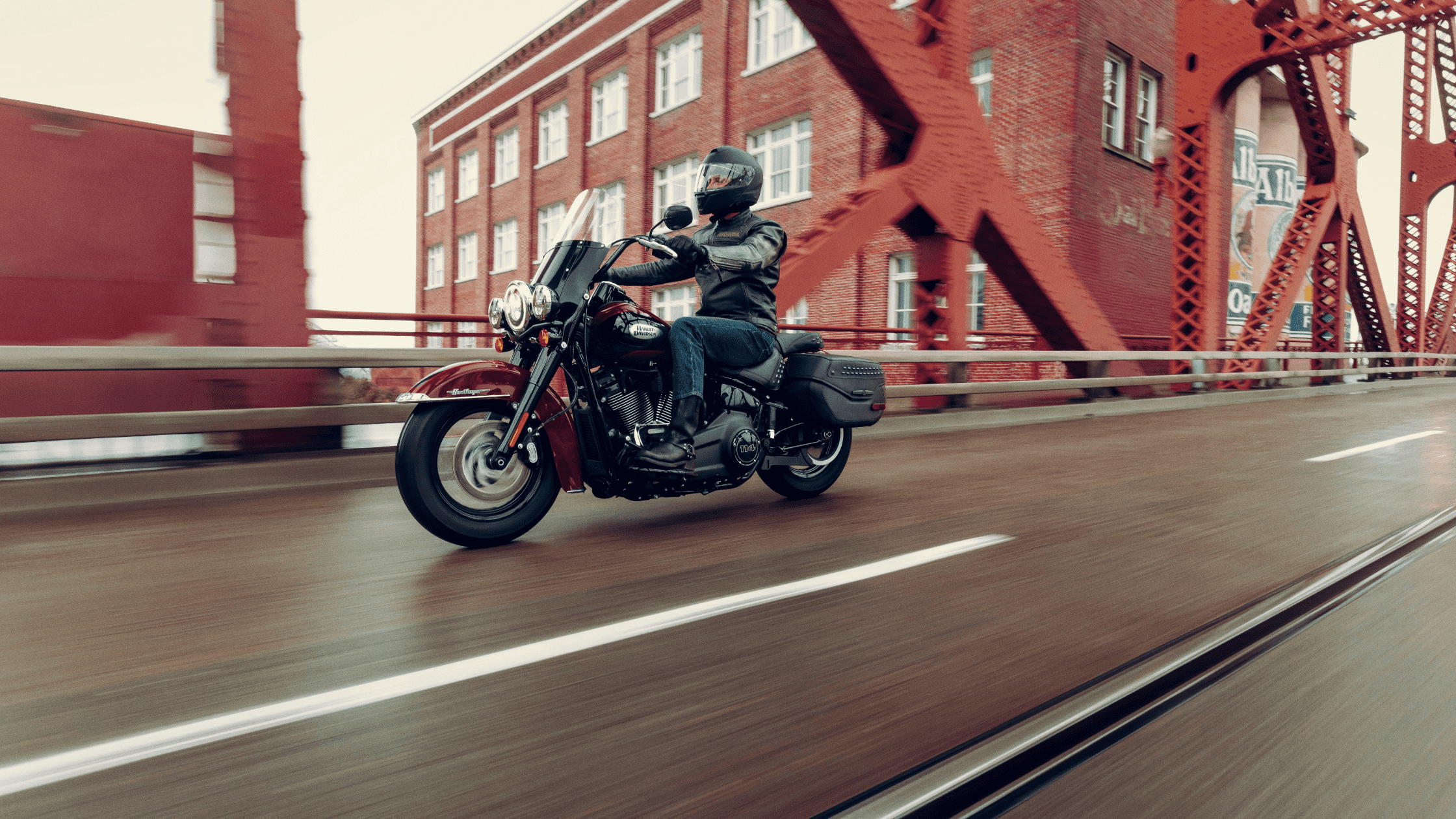 A rider cruising on a 2024 Harley-Davidson® Heritage Classic across a red steel bridge with brick buildings in the background