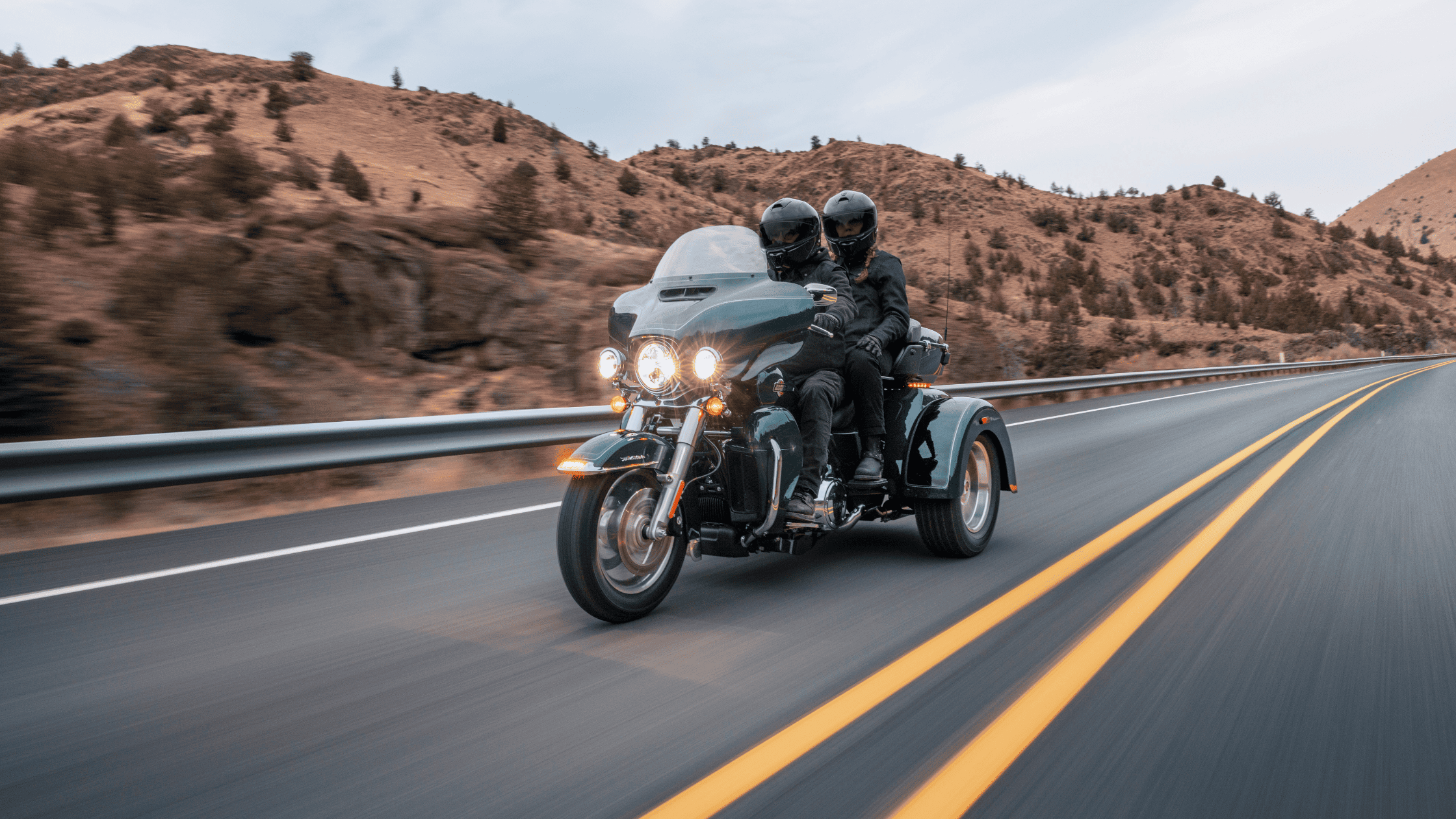 Two riders cruising on a Harley-Davidson Tri Glide® Ultra along a scenic highway, surrounded by rolling desert hills.