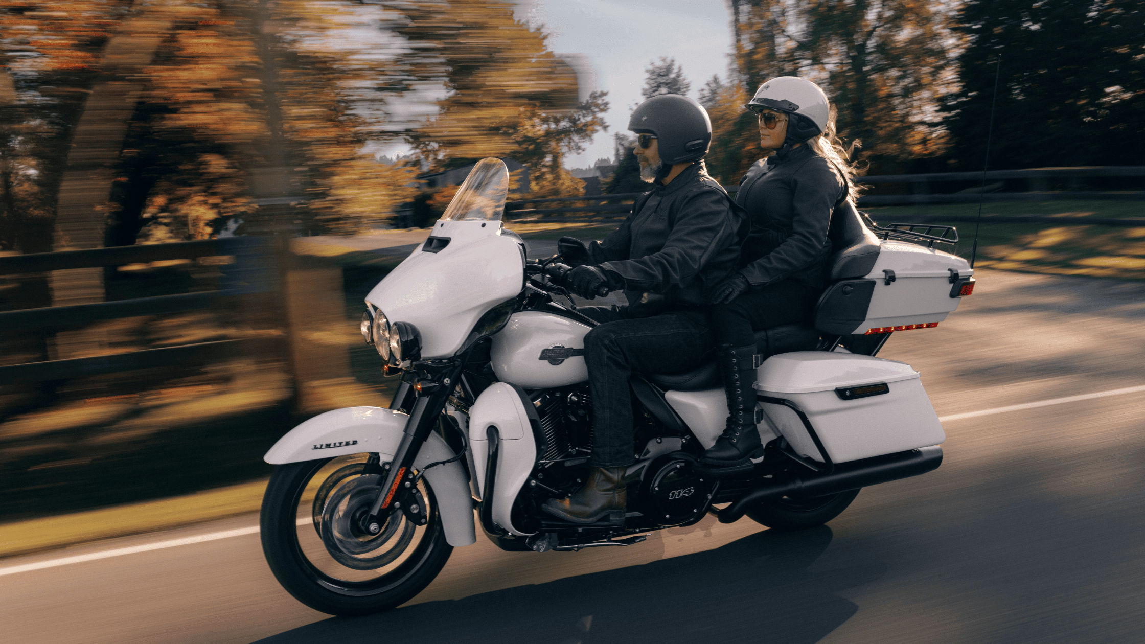 A rider and passenger cruising on a 2024 Harley-Davidson® Ultra Limited, riding down a tree-lined road in autumn