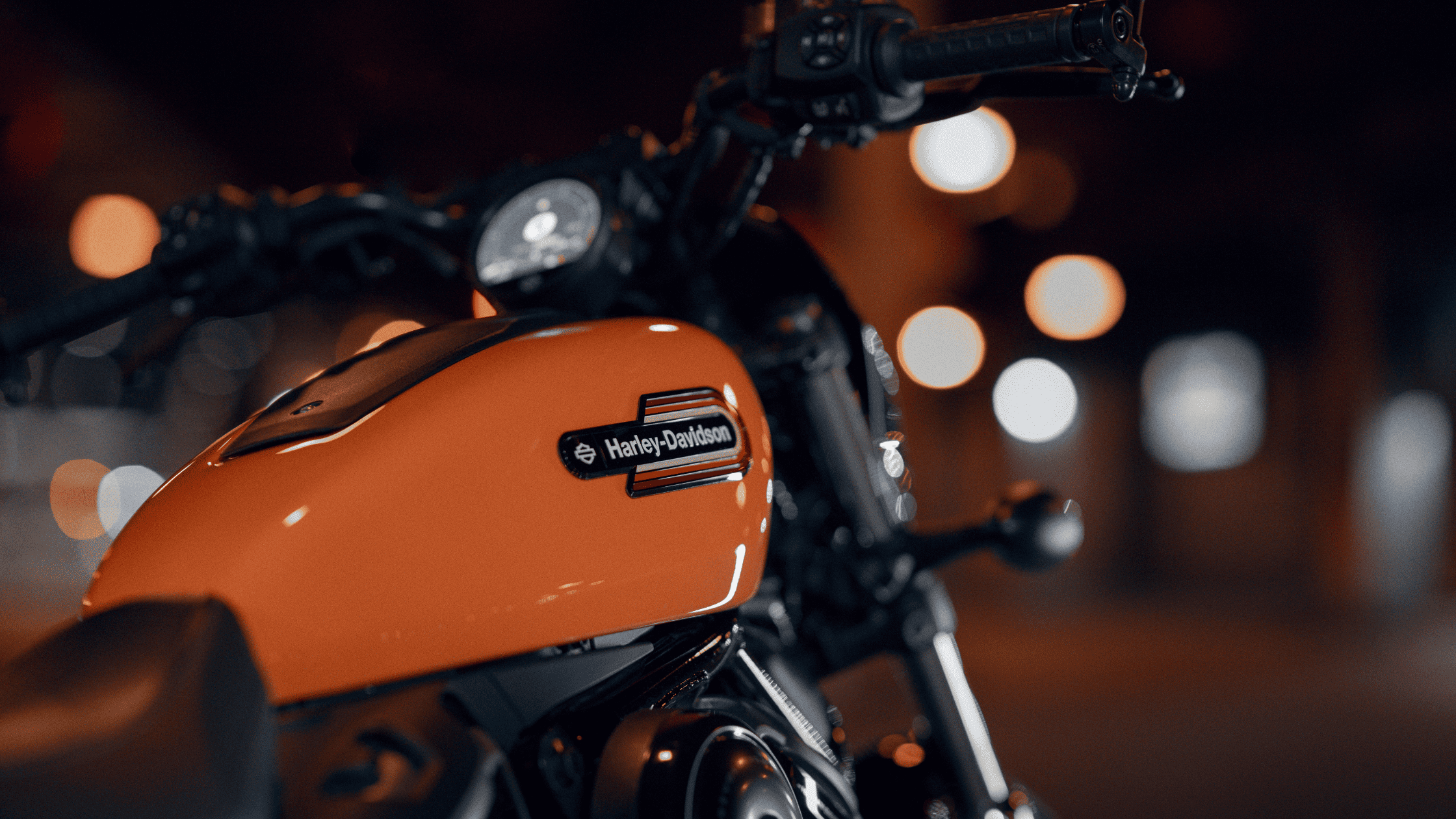 Close-up of an orange Harley-Davidson Nightster® Special fuel tank, with blurred city lights in the background creating a moody atmosphere.