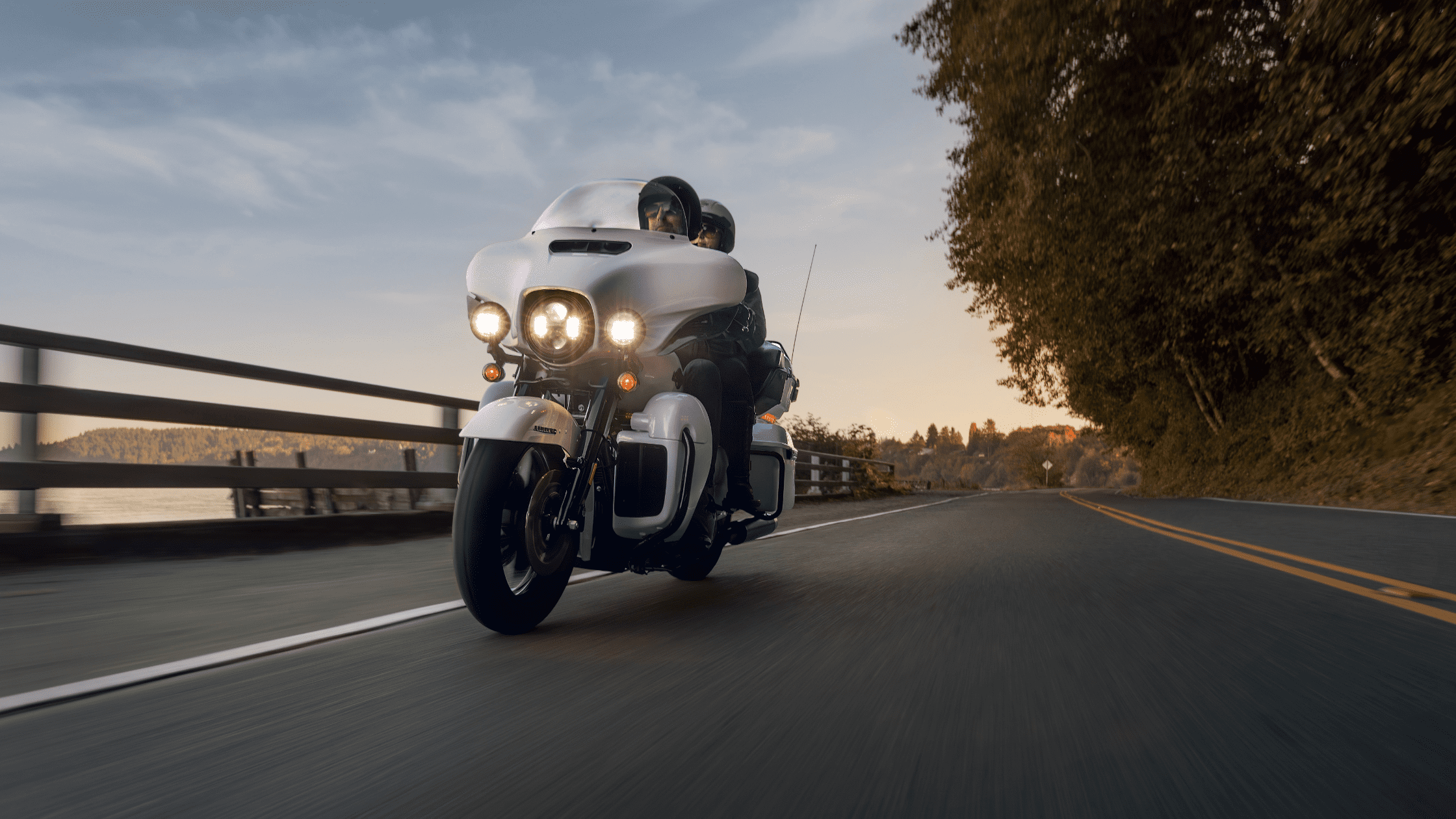 Two riders on a Harley-Davidson Ultra Limited® cruising down a scenic lakeside road at sunset, with trees and water lining the background.