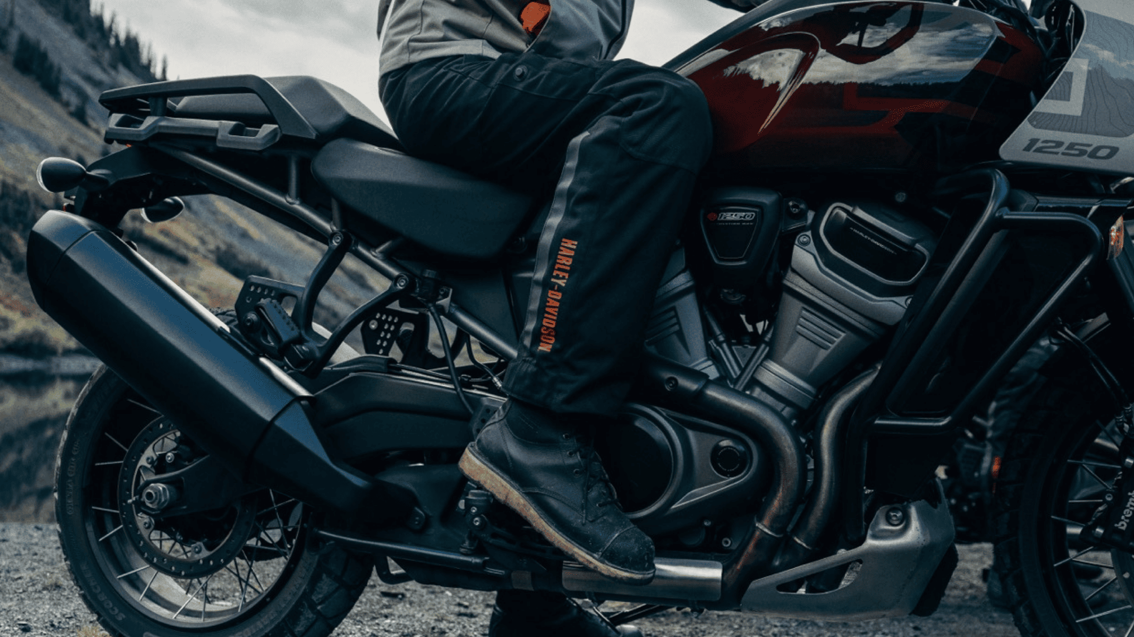 Close-up of a rider’s leg on a Harley-Davidson Pan America® 1250 Special, showing detailed gear and motorcycle components, with a scenic mountain backdrop.
