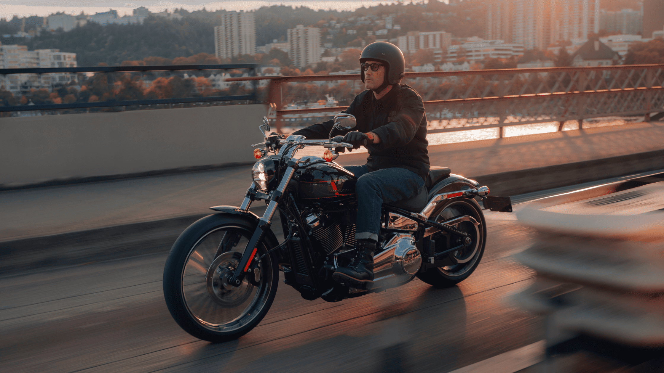 Rider cruising on a Harley-Davidson Breakout® 117 across a city bridge at sunset, with an urban skyline and tree-covered hills in the background.