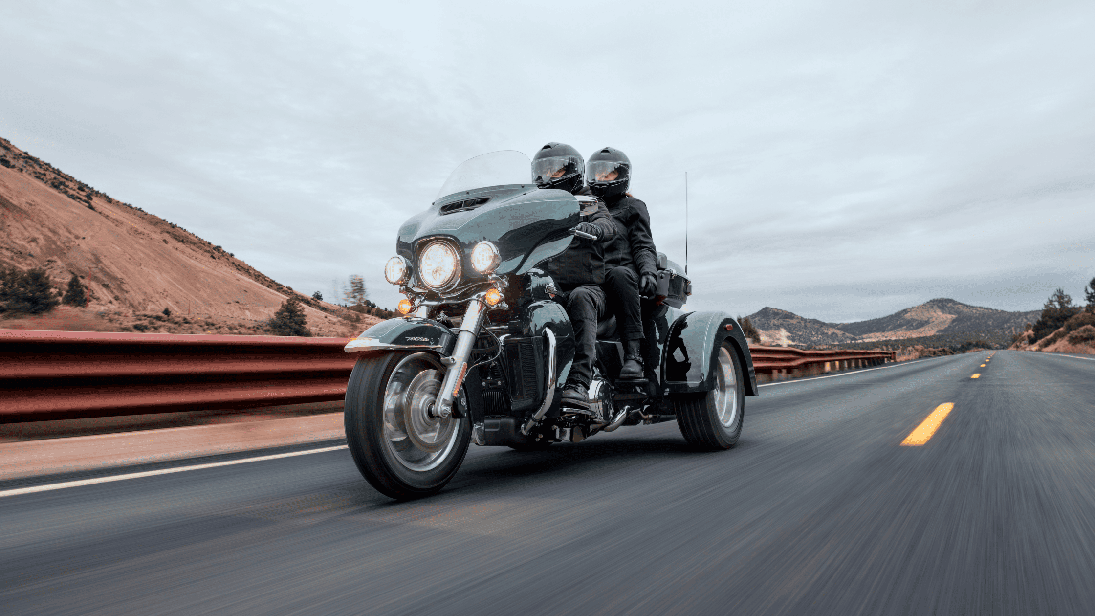 Two riders on a Harley-Davidson Tri Glide® Ultra cruising down a scenic highway, with rolling hills and overcast skies in the background.