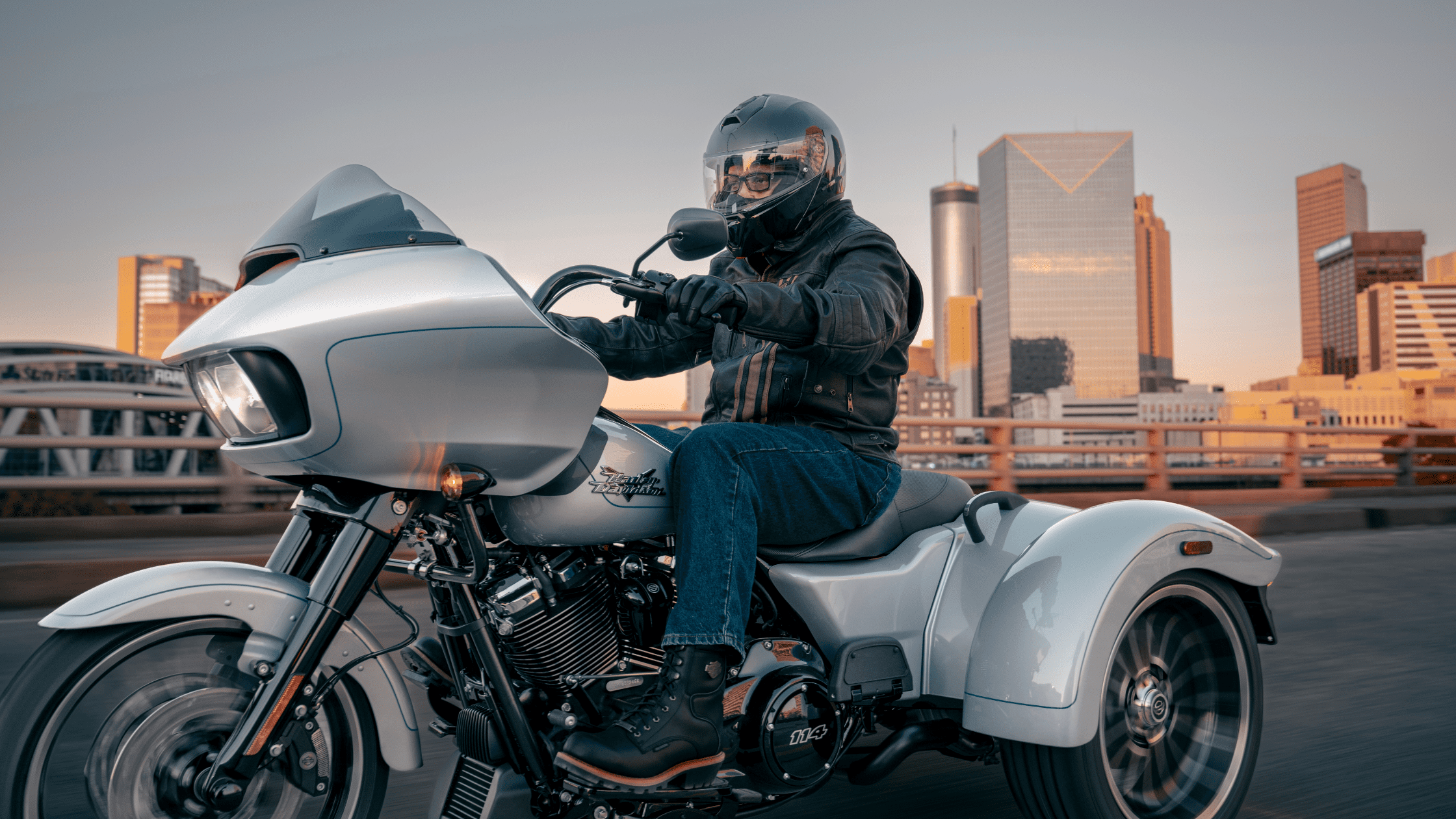 Rider cruising on a Harley-Davidson Road Glide® 3 through a city at sunset, with modern skyscrapers in the background.