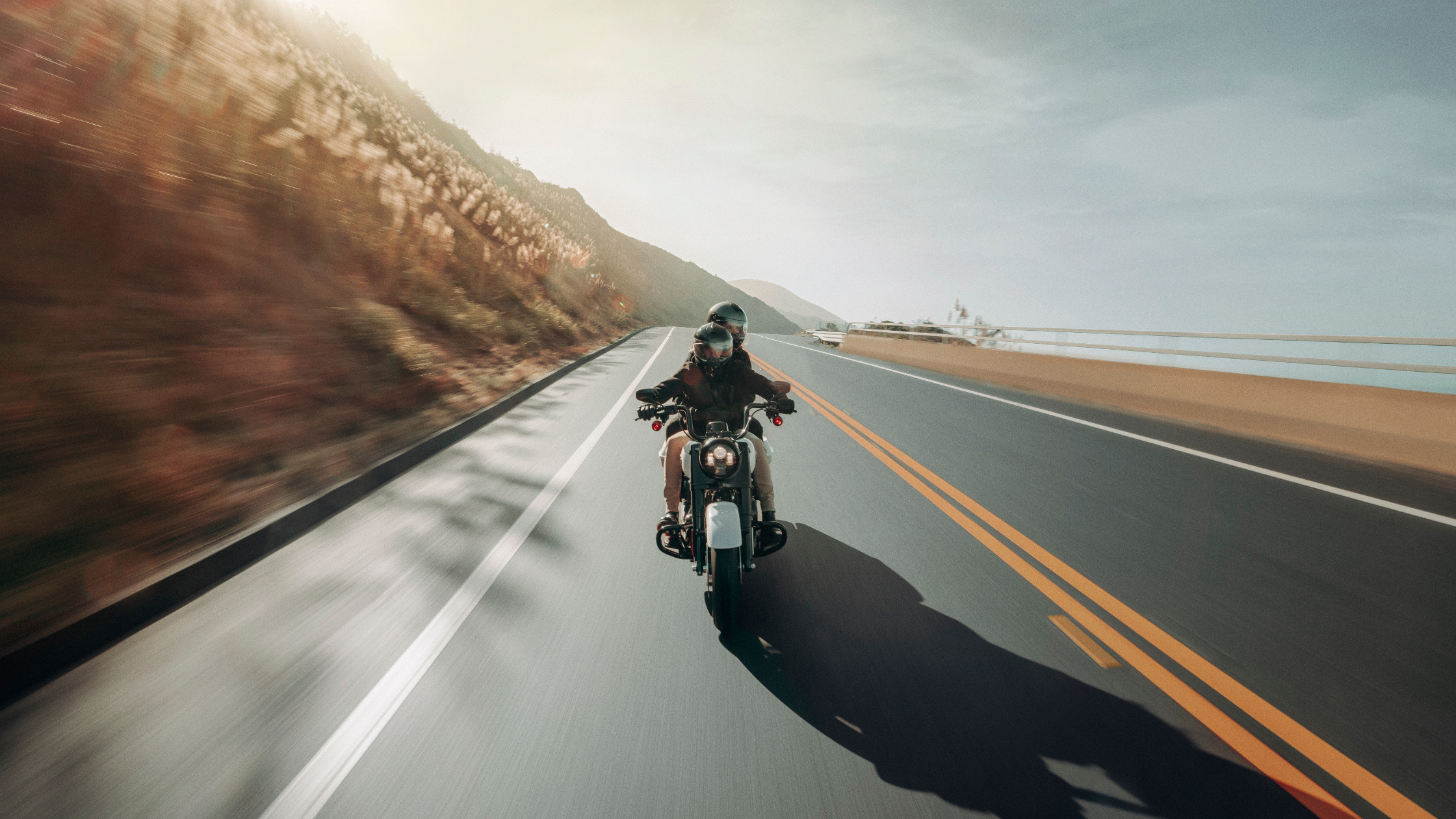 Two riders cruise along a scenic highway on a motorcycle, with the sun shining over a mountainous landscape and ocean views in the distance.
