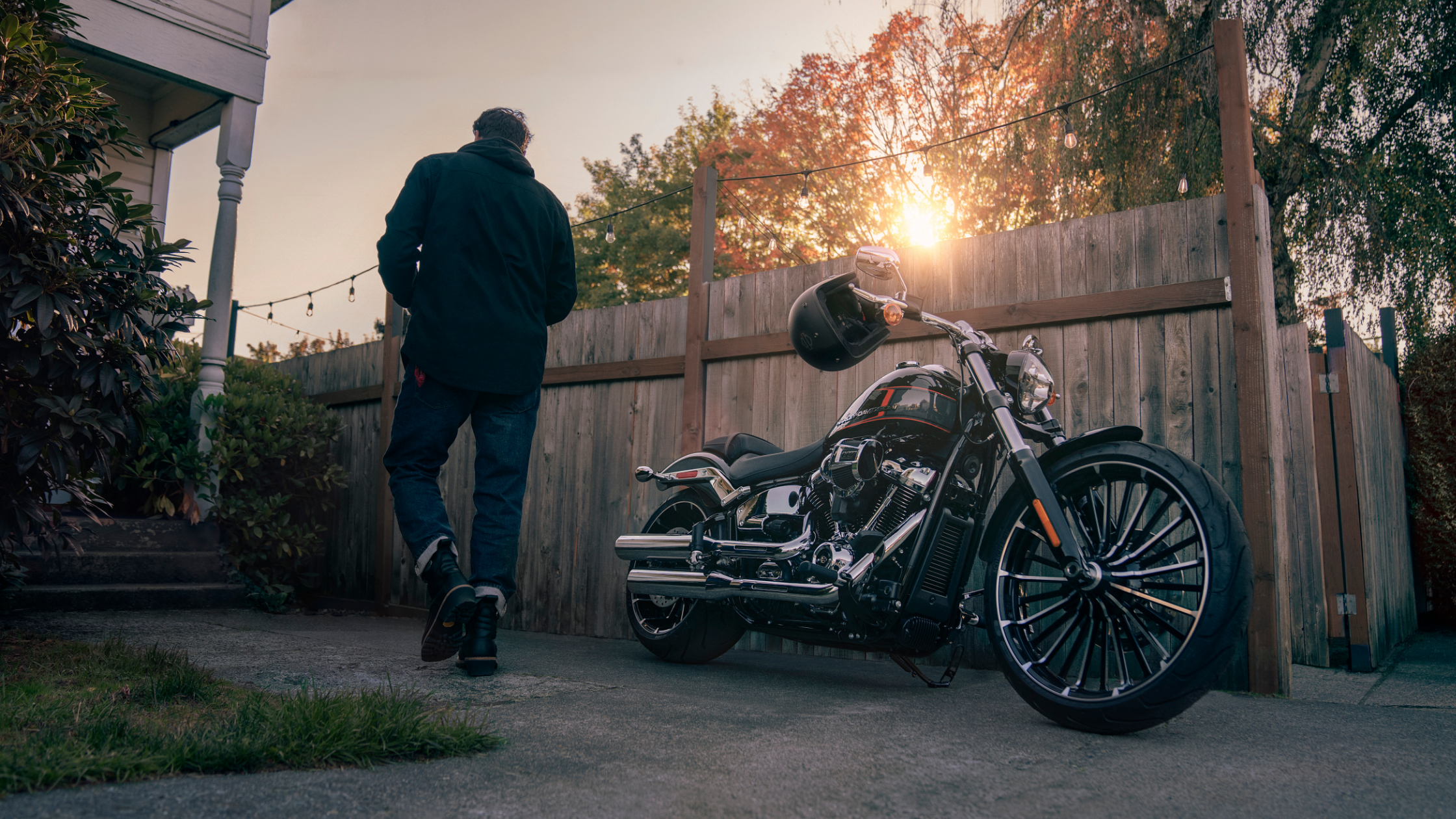 A man walks towards a wooden gate at sunset, leaving behind a parked Harley-Davidson® motorcycle with a helmet resting on the handlebar.