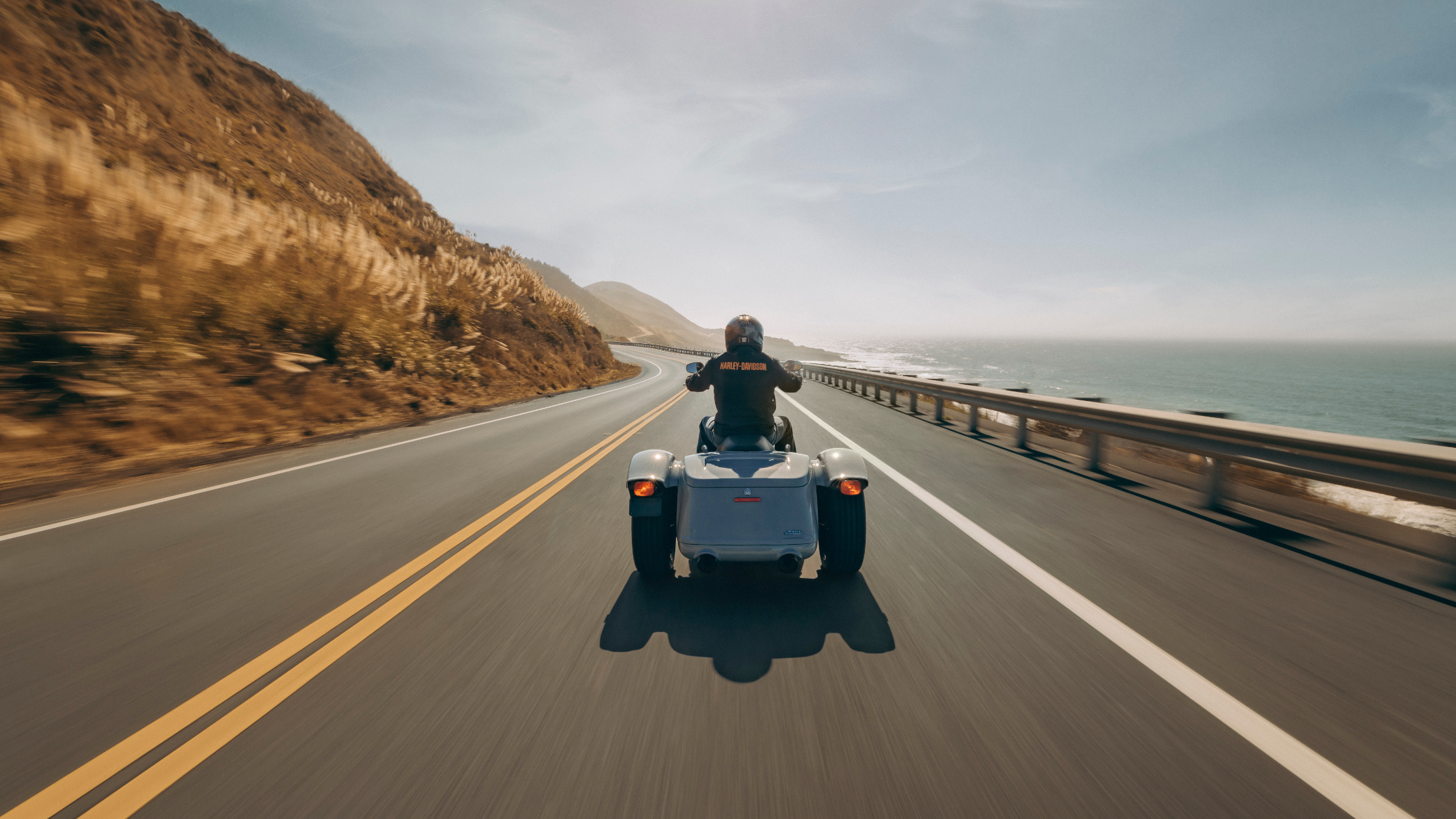 A rider on a Harley-Davidson® trike cruises along a coastal highway, with the ocean to the right and hills to the left under a bright sky.