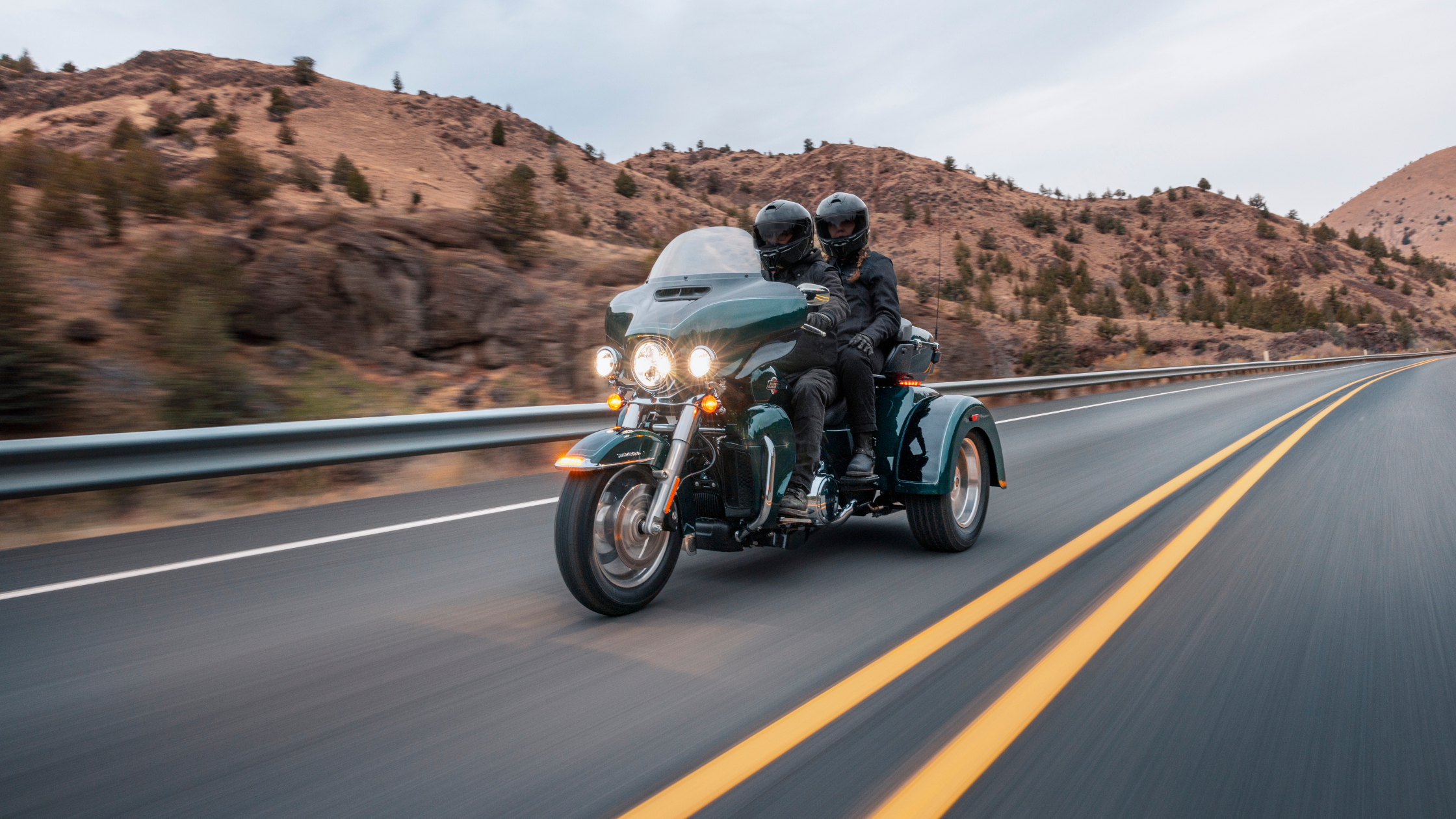 Two riders on a Harley-Davidson® trike cruise down a scenic highway through a dry, hilly landscape.