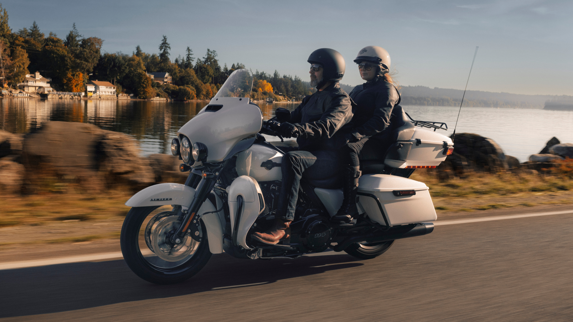 Two riders on a Harley-Davidson® motorcycle cruise along a lakeside road on a sunny day, with trees and houses reflecting on the calm water.