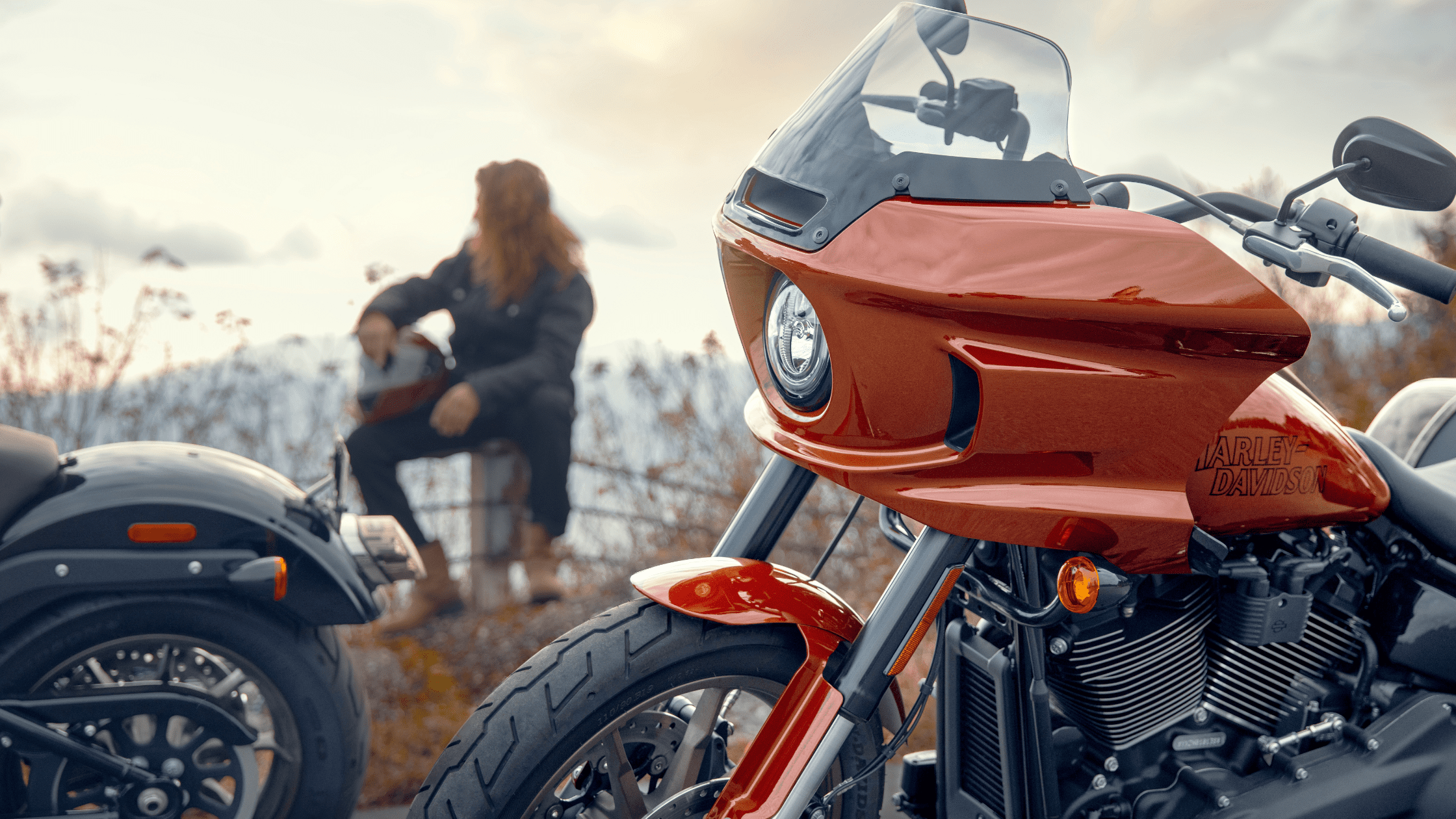 A close-up of a 2024 Harley-Davidson® Low Rider® ST with a rider sitting in the background, gazing out over a scenic landscape