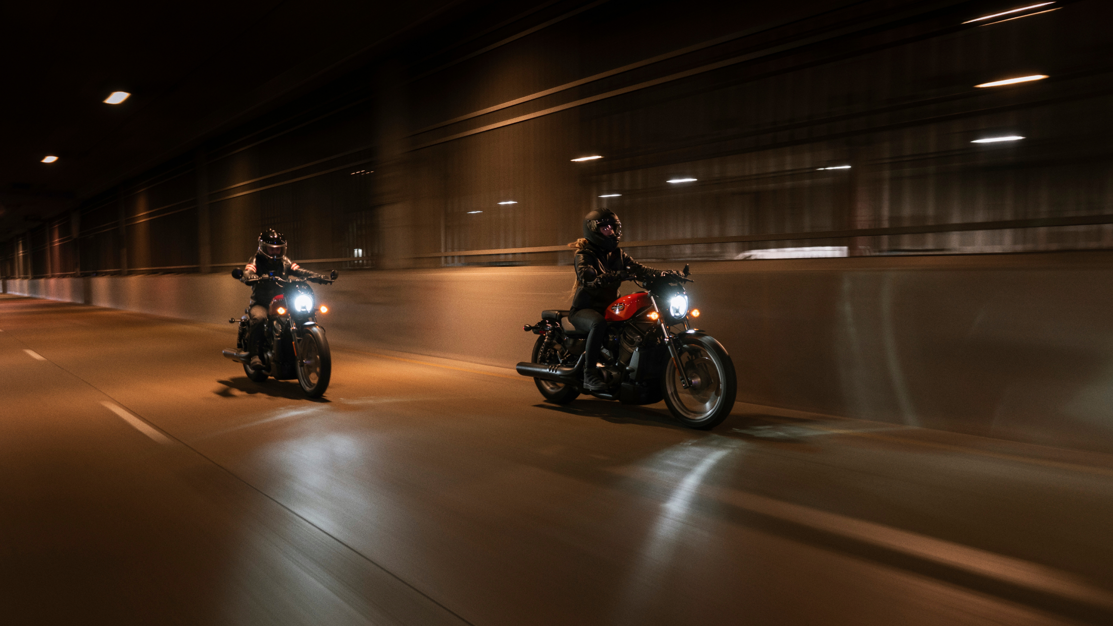 Two riders on Harley-Davidson® motorcycles cruise through a dimly lit tunnel at night.