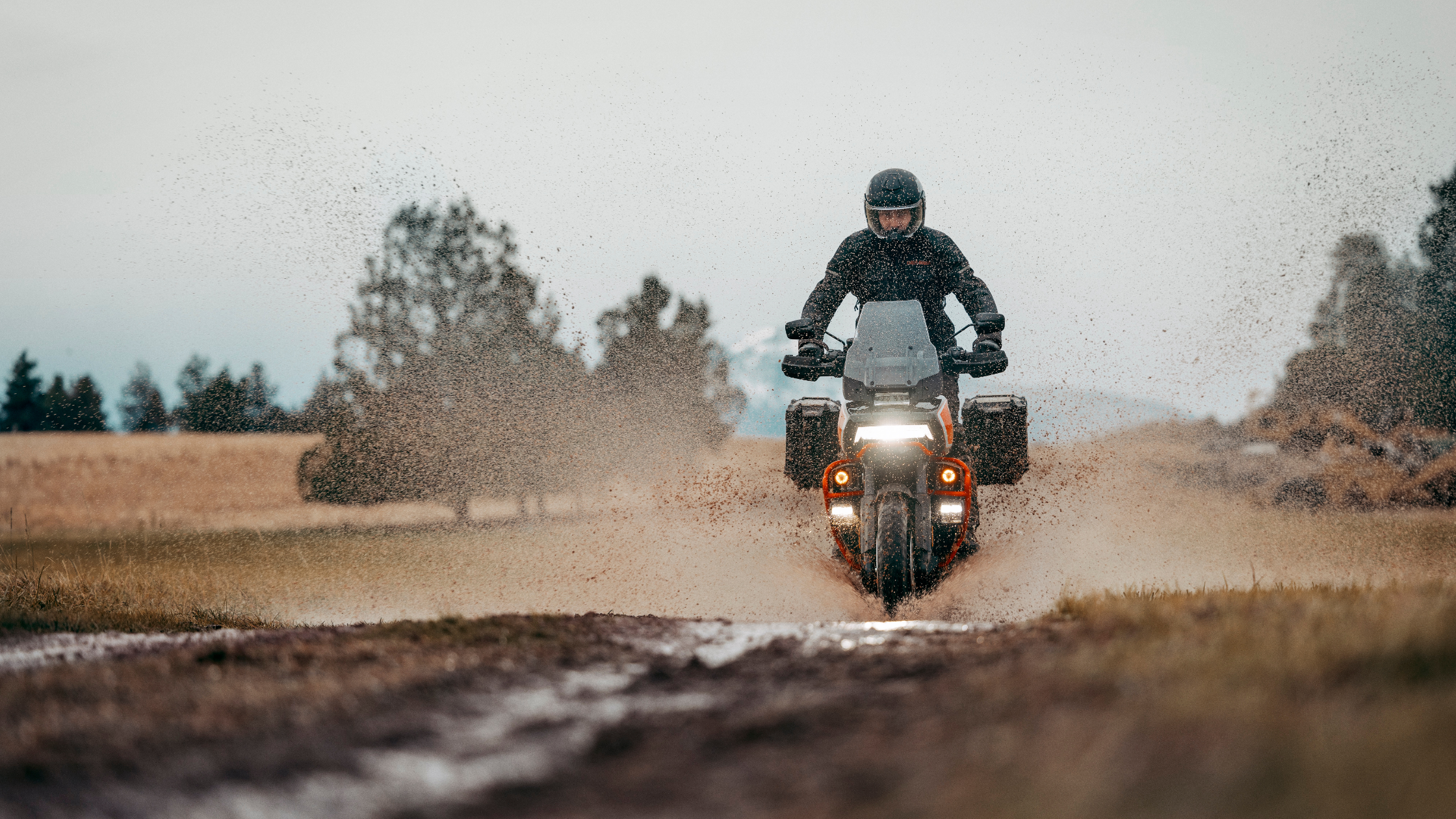 A rider on a Harley-Davidson® adventure motorcycle splashes through a muddy trail, kicking up dirt and water in an open rural landscape.