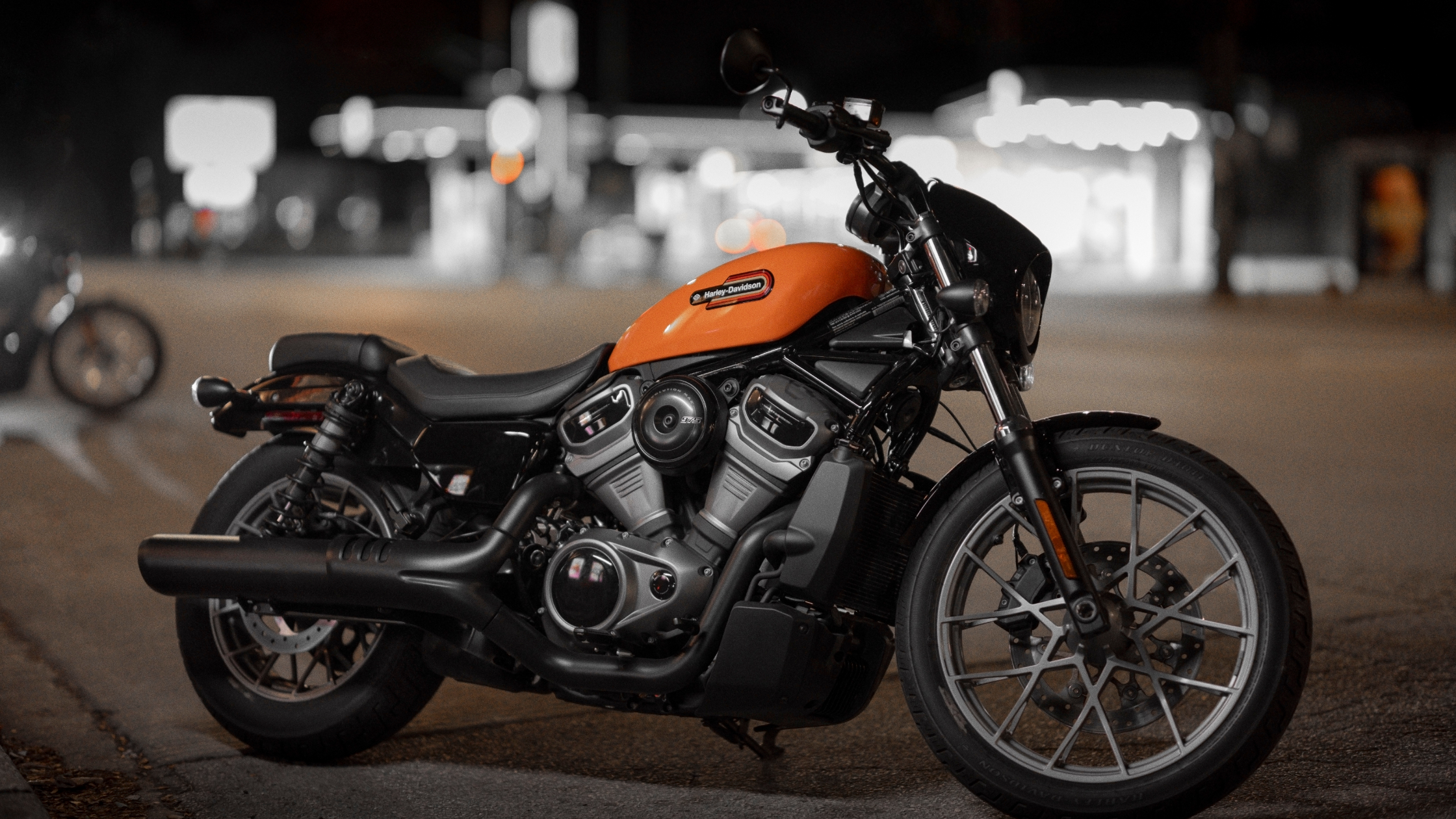 A 2024 Harley-Davidson® Nightster® in orange is parked under streetlights at night, with a blurred city background.