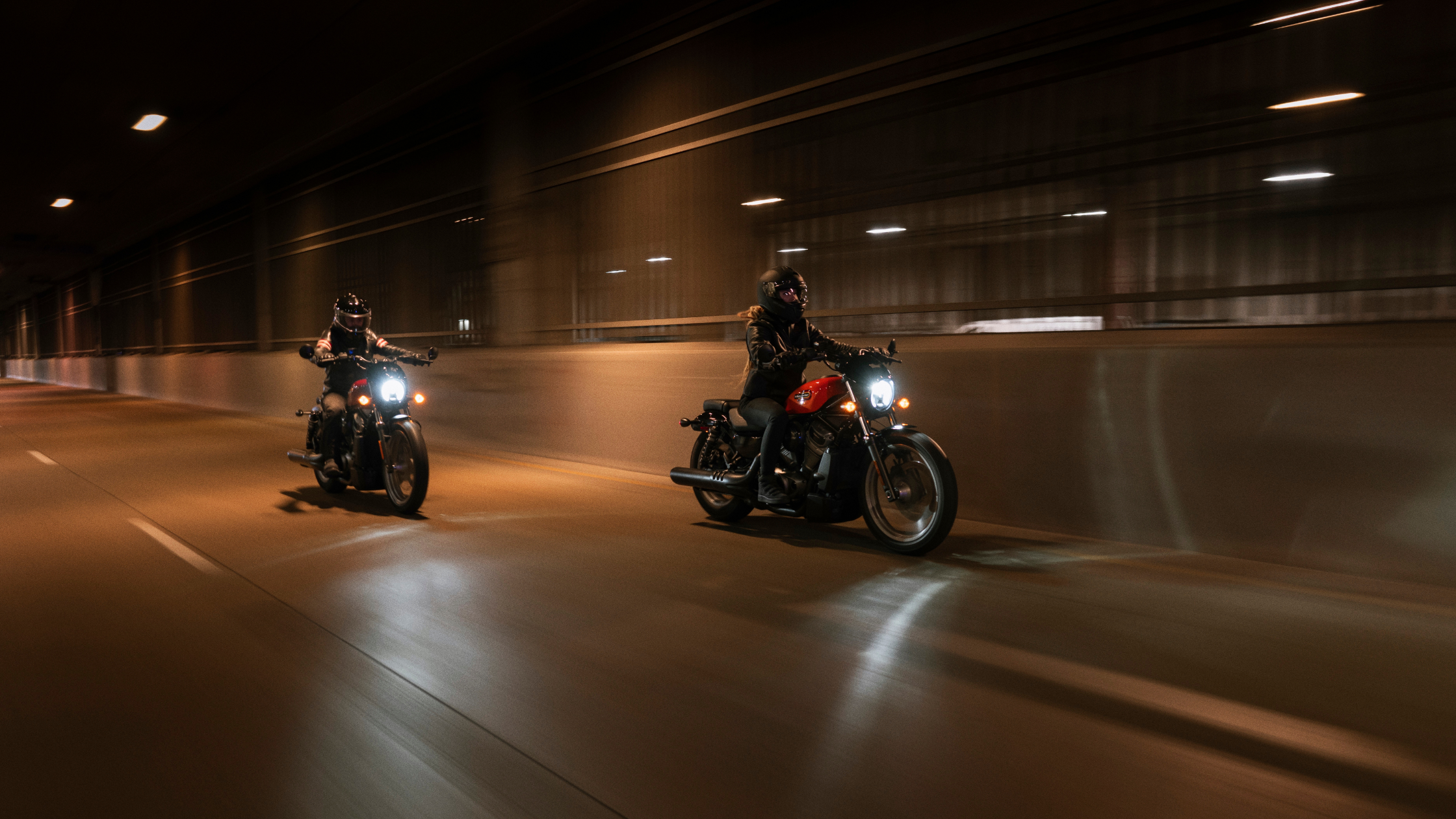Two riders on Harley-Davidson® motorcycles ride through a dimly lit tunnel at night, their headlights cutting through the darkness.
