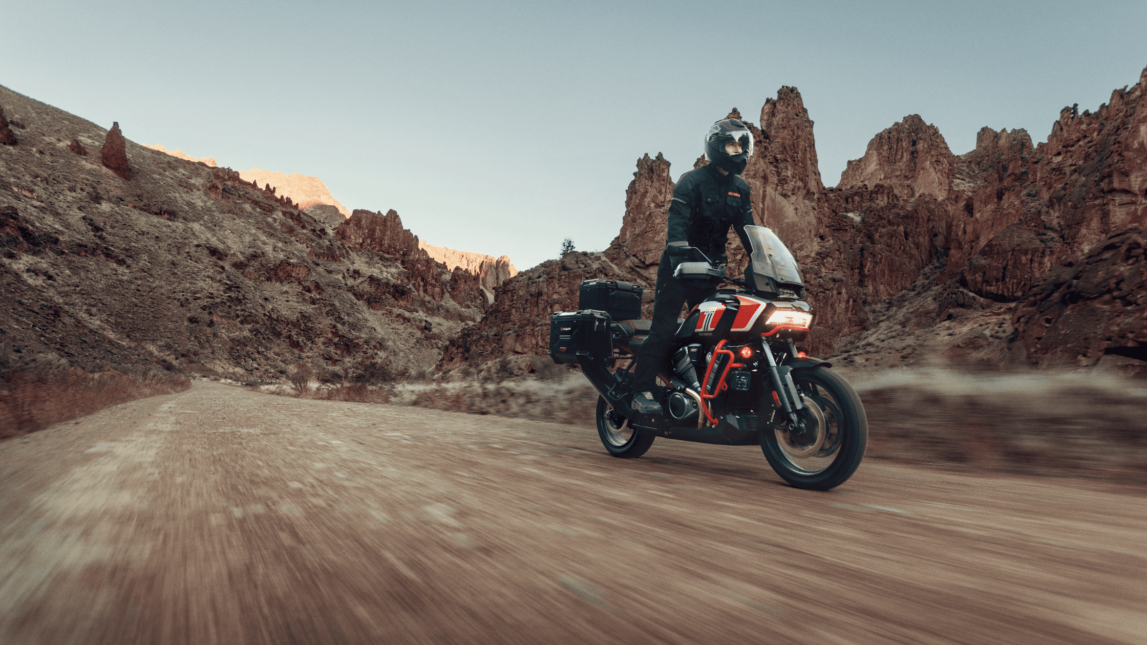 Rider on a Harley-Davidson® CVO™ Pan America® motorcycle driving through a rugged desert landscape with rocky cliffs in the background.