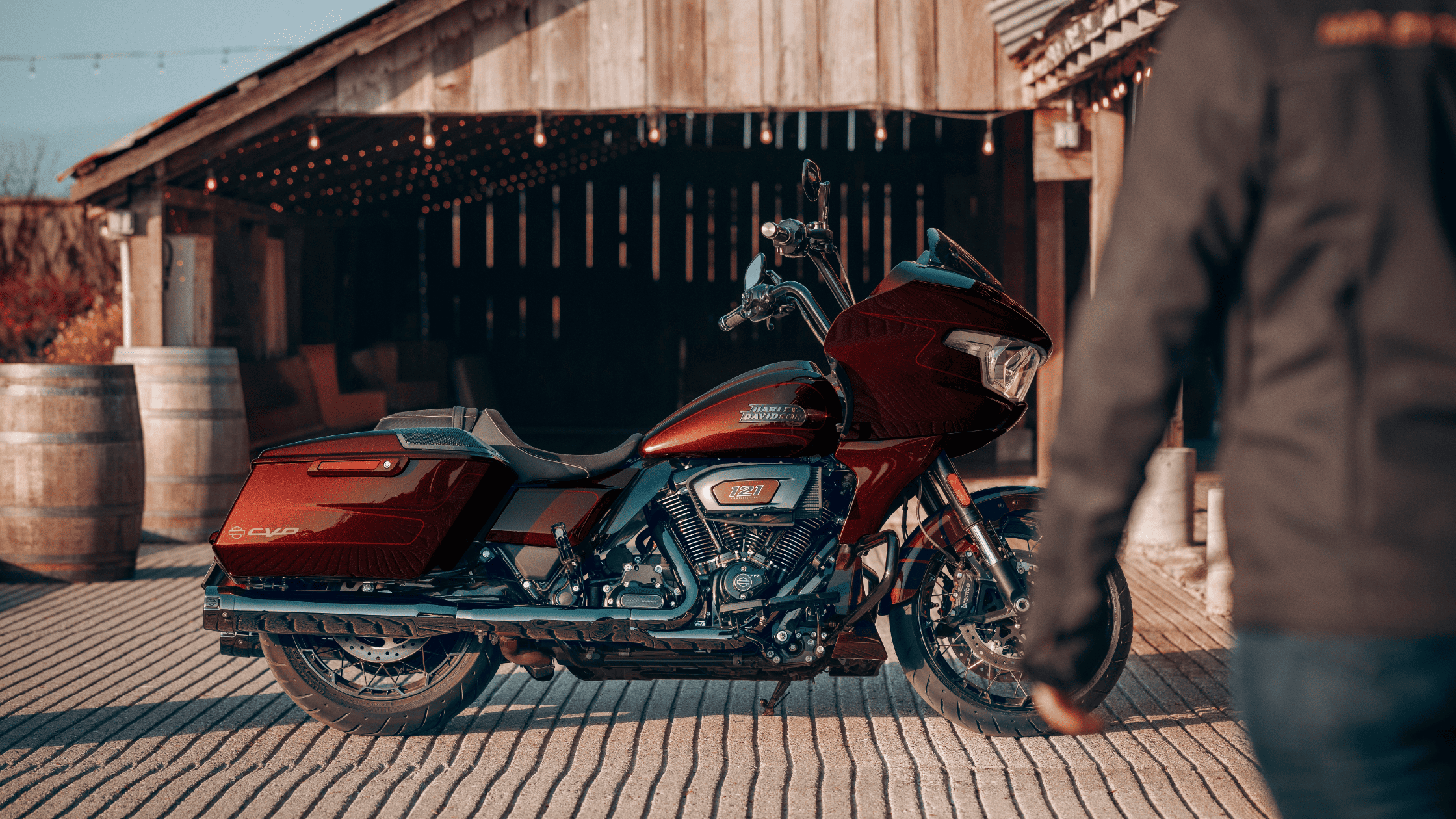 2024 Harley-Davidson CVO Road Glide in deep red, parked in front of a rustic barn with wooden barrels, as a rider approaches from the side.