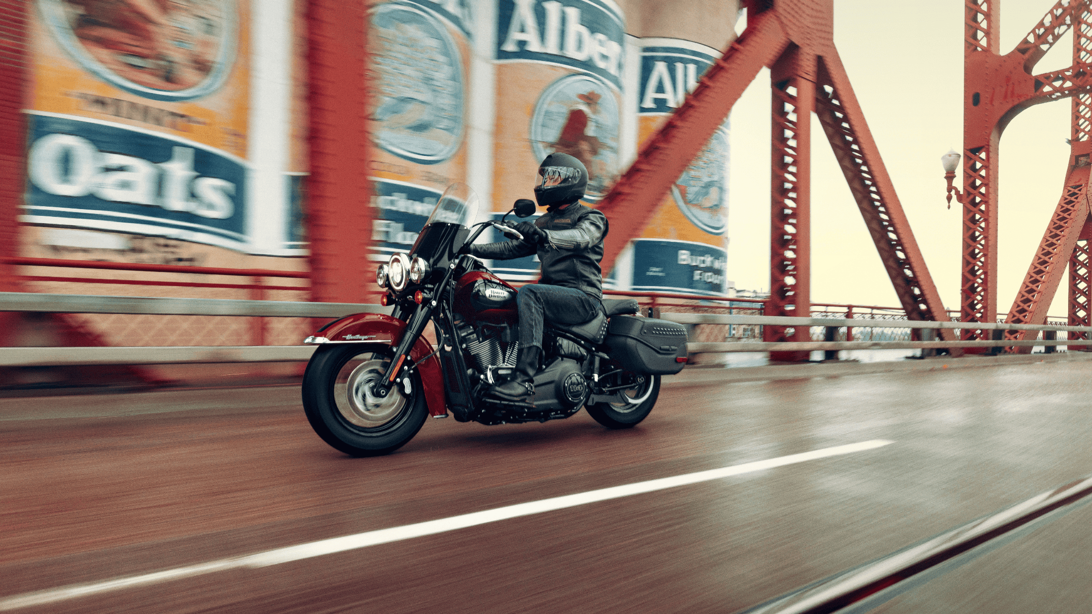 Rider on a 2024 Harley-Davidson Heritage Classic 114 motorcycle cruising over a red steel bridge with vintage billboard ads in the background.