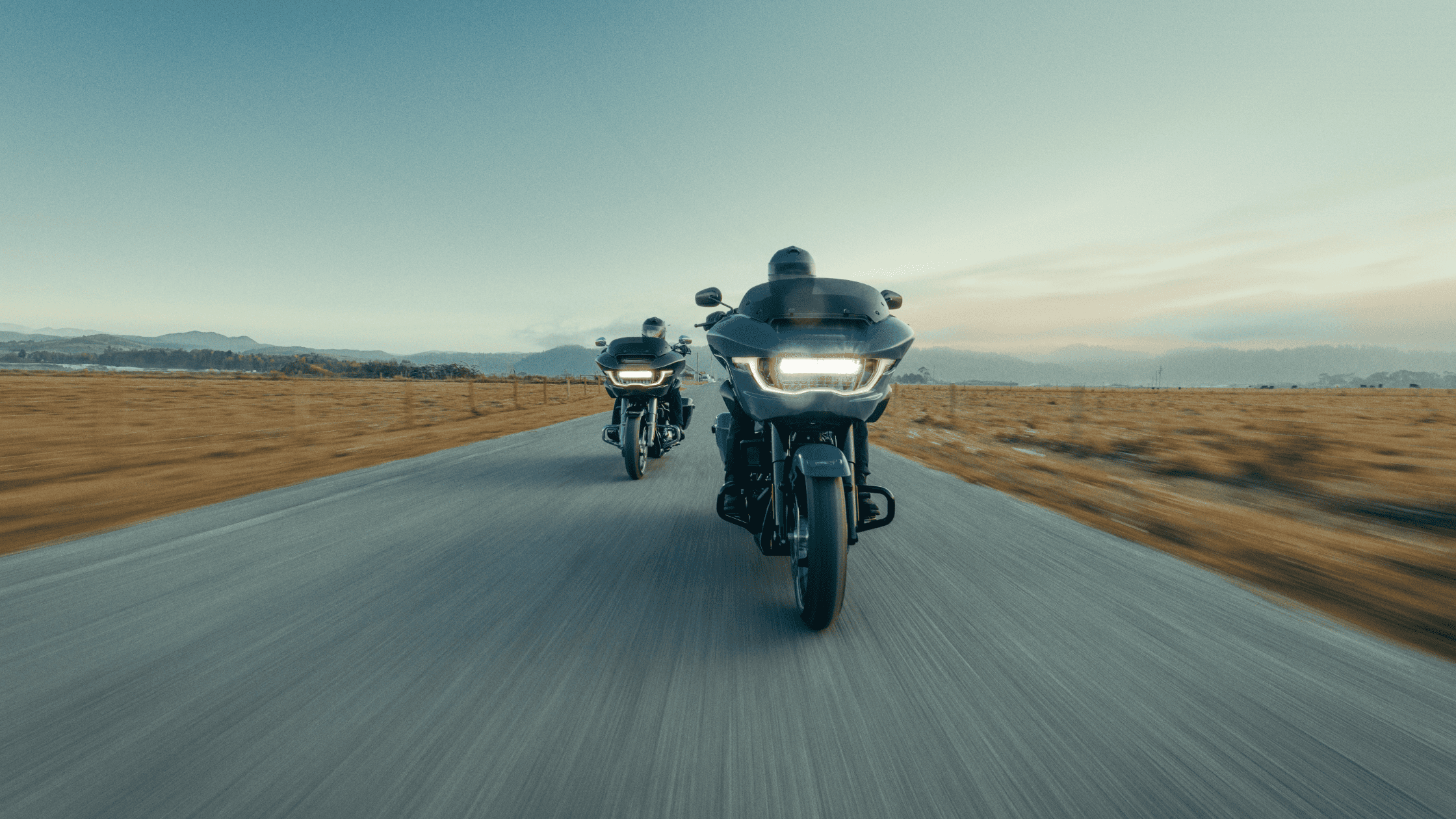 Two riders on 2024 Harley-Davidson Road Glide motorcycles cruising down a straight, open road with scenic mountain views in the background.