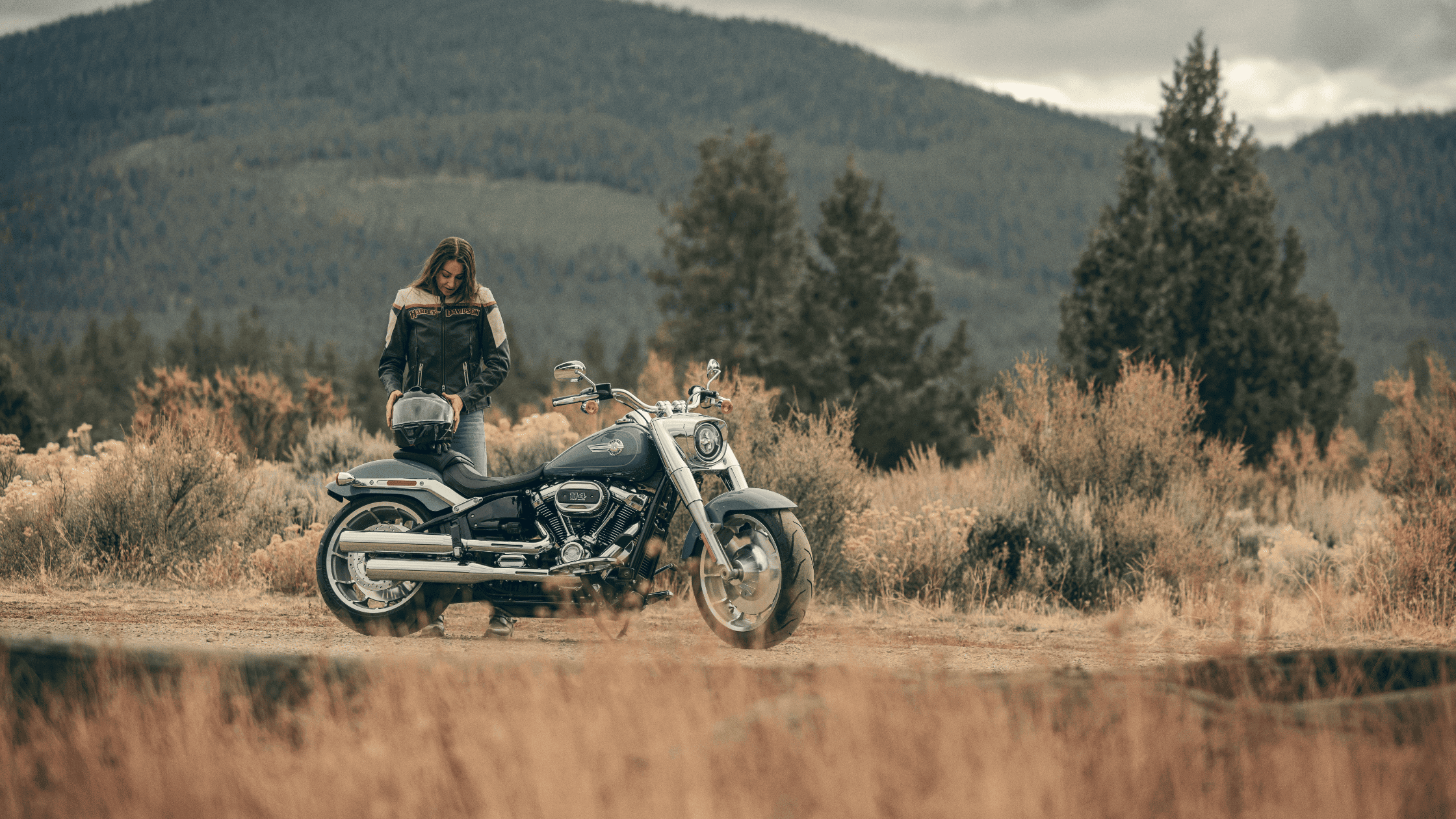 Rider standing next to a 2024 Harley-Davidson Fat Boy 114 motorcycle in a scenic outdoor landscape, with mountains and trees in the background.