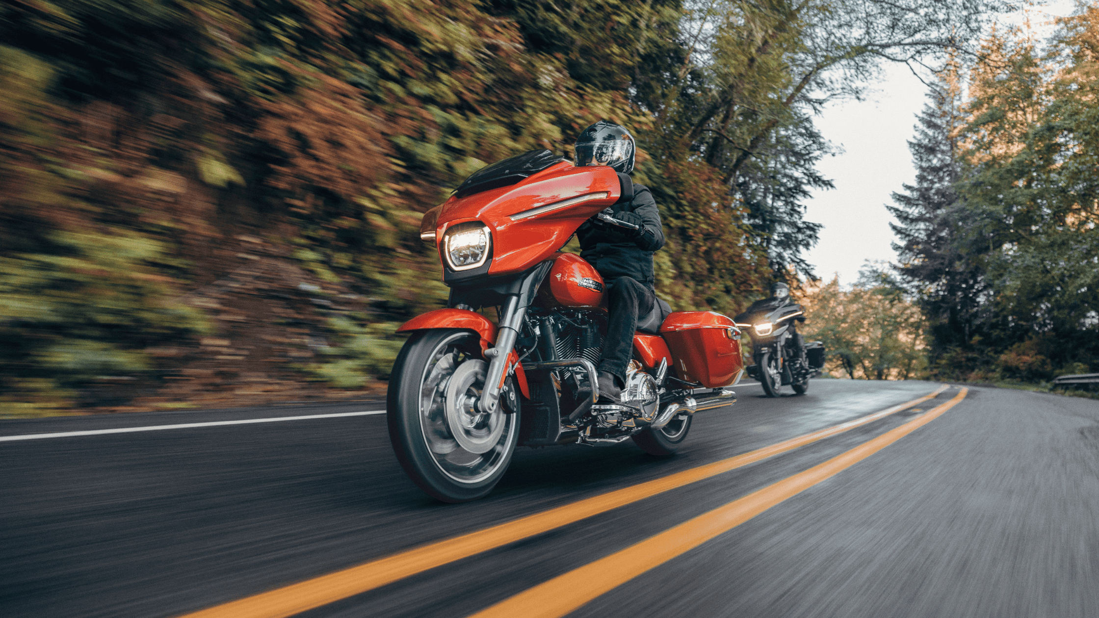 Rider cruising on a 2024 Harley-Davidson Street Glide in vibrant orange, leading a scenic ride through a tree-lined mountain road.