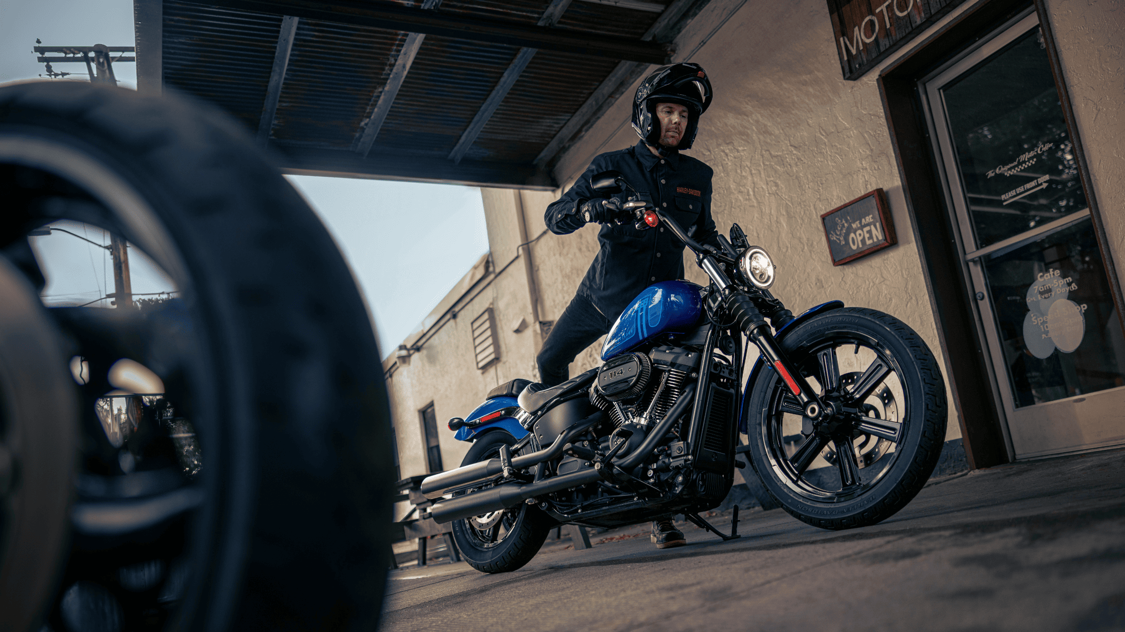 Rider preparing to set off on a 2024 Harley-Davidson Street Bob 114 motorcycle in vivid blue, parked outside a vintage-style café.