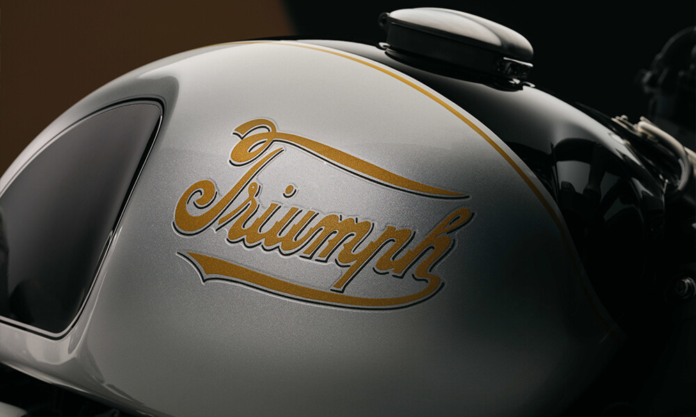 Close up side on image of a Triumphs fuel tank in aluminium silver featuring the gold script Triumph logo