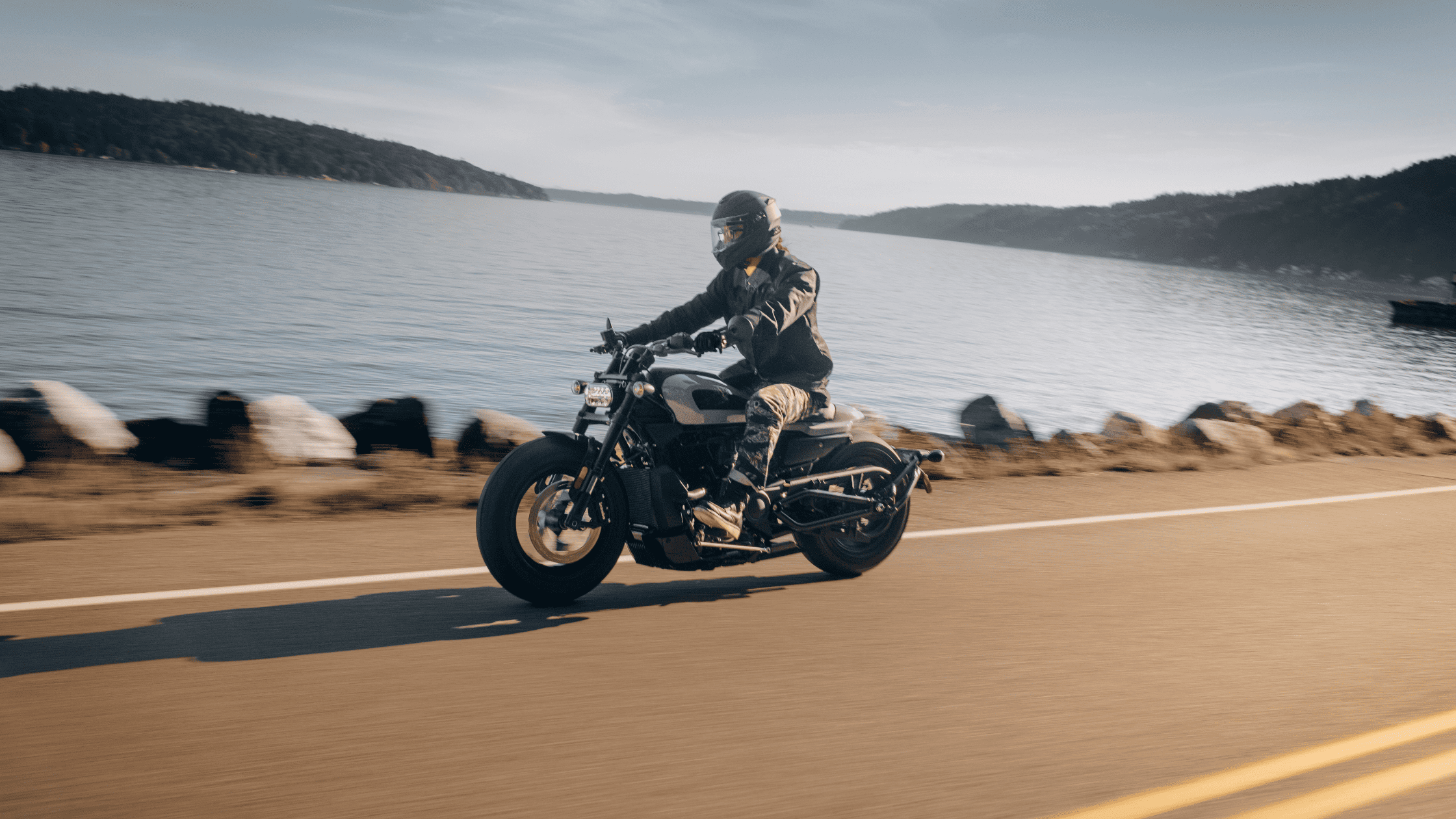 Motorcyclist riding along a serene lakeside road in daylight.